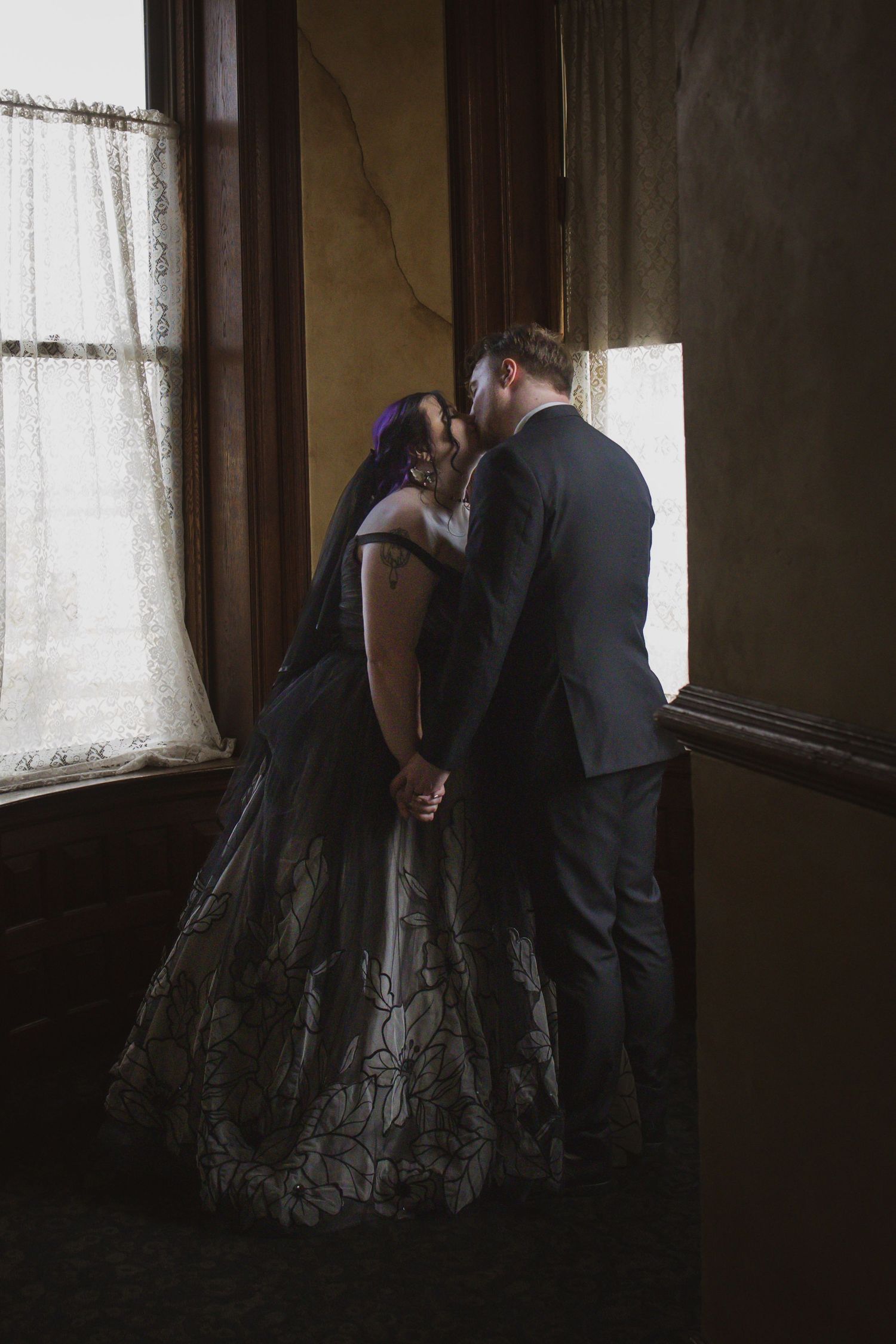 A couple in wedding attire share a tender moment by a window in moody lighting.