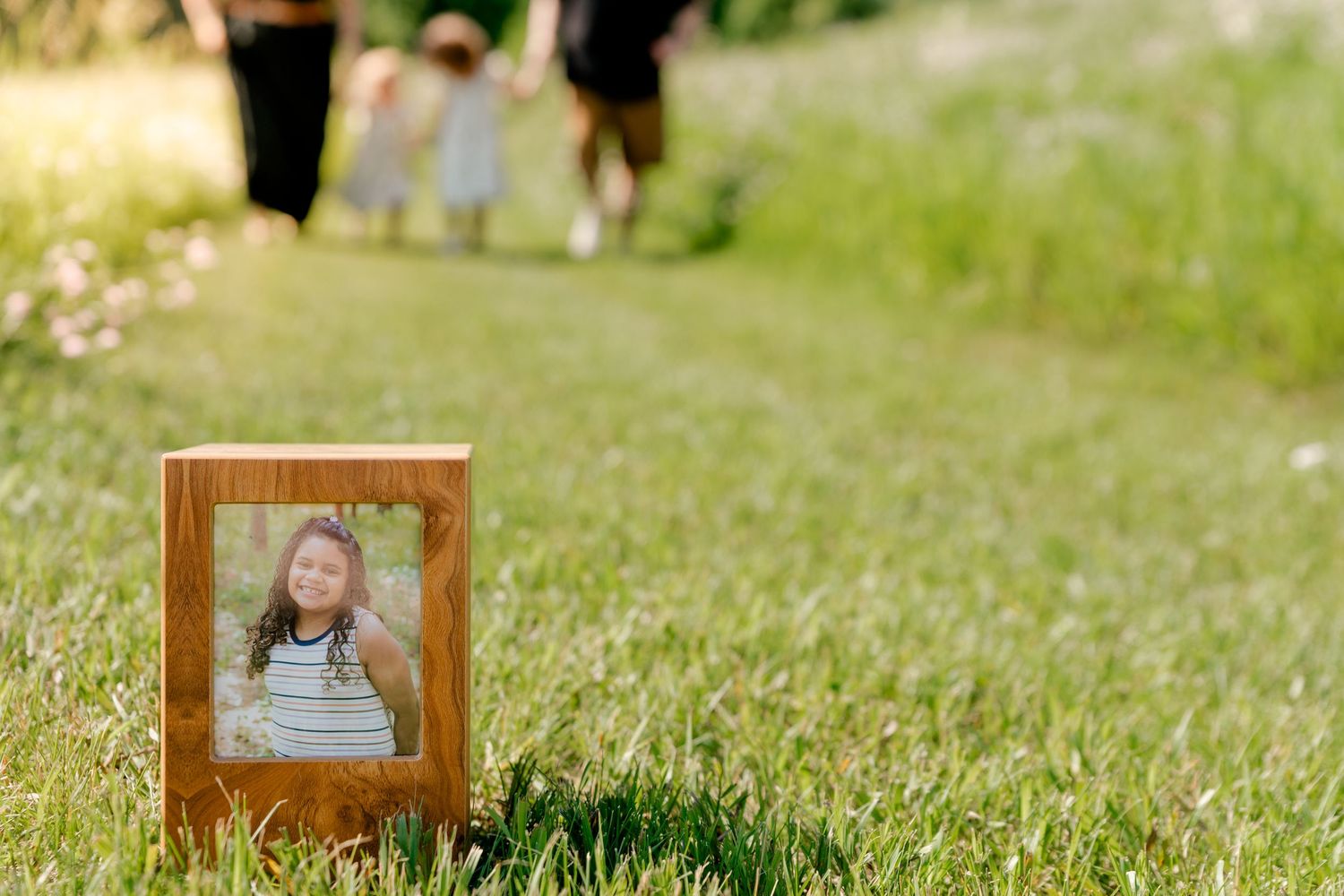 A framed portrait photograph sits in green grass with blurred figures walking in the background.