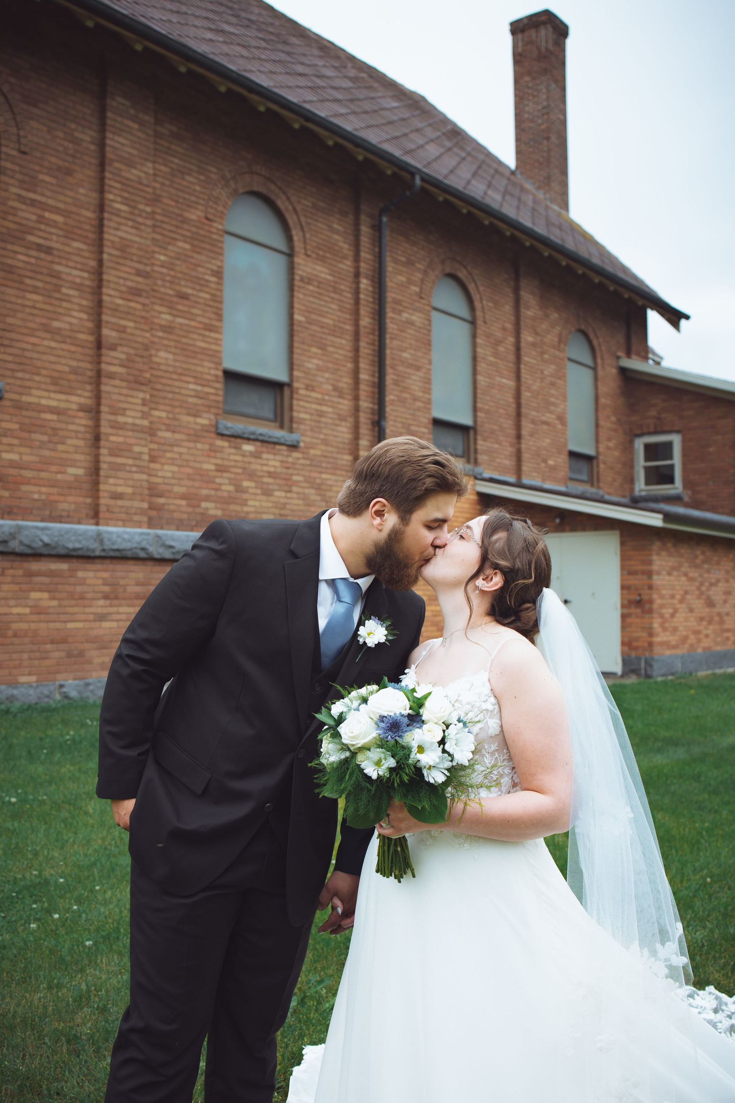 A wedding couple shares a romantic moment outside a brick church with stained glass windows and sloping rooflines.