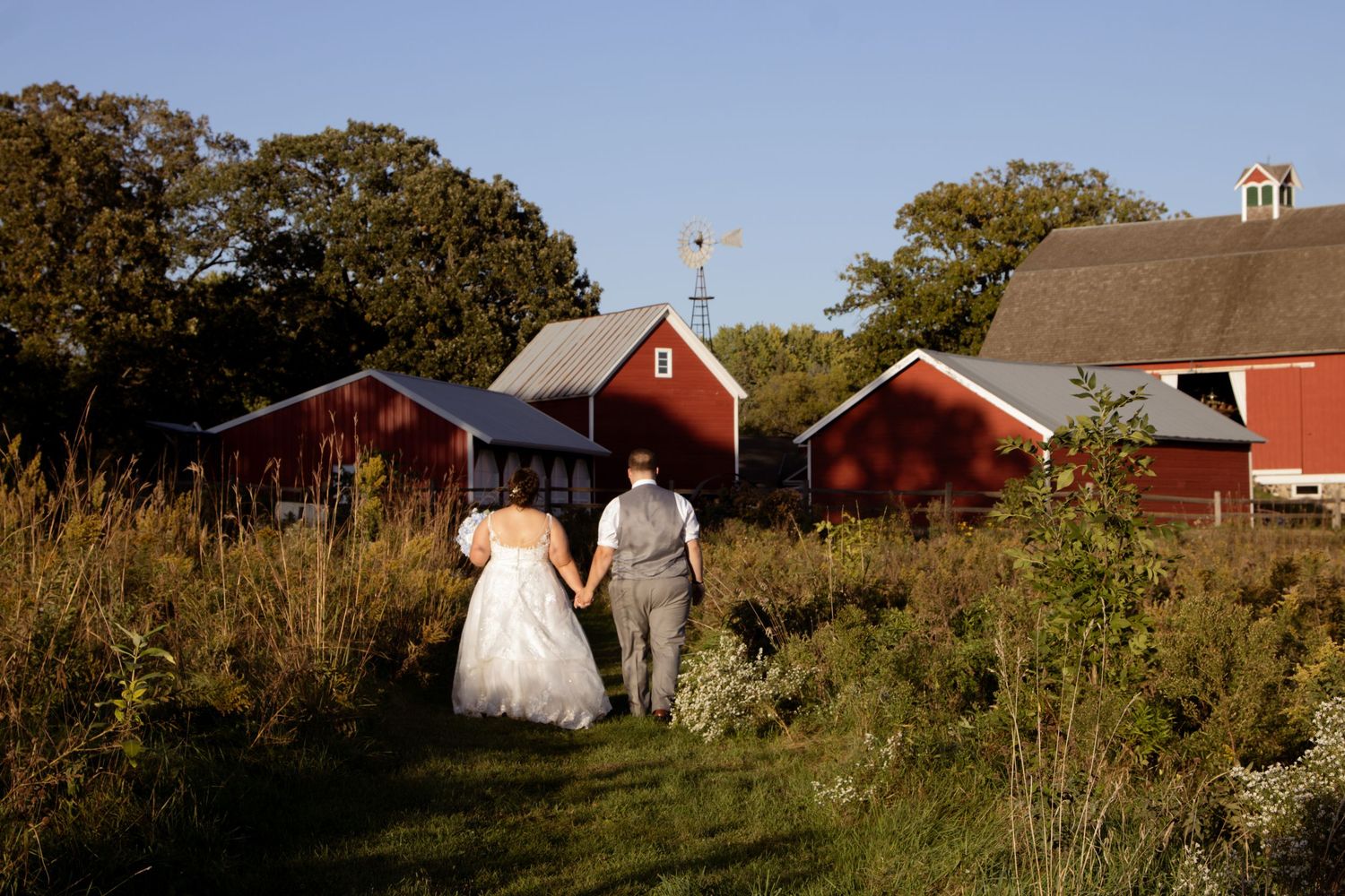 Two figures in wedding attire walk along a path near rustic red barns and trees during golden hour.