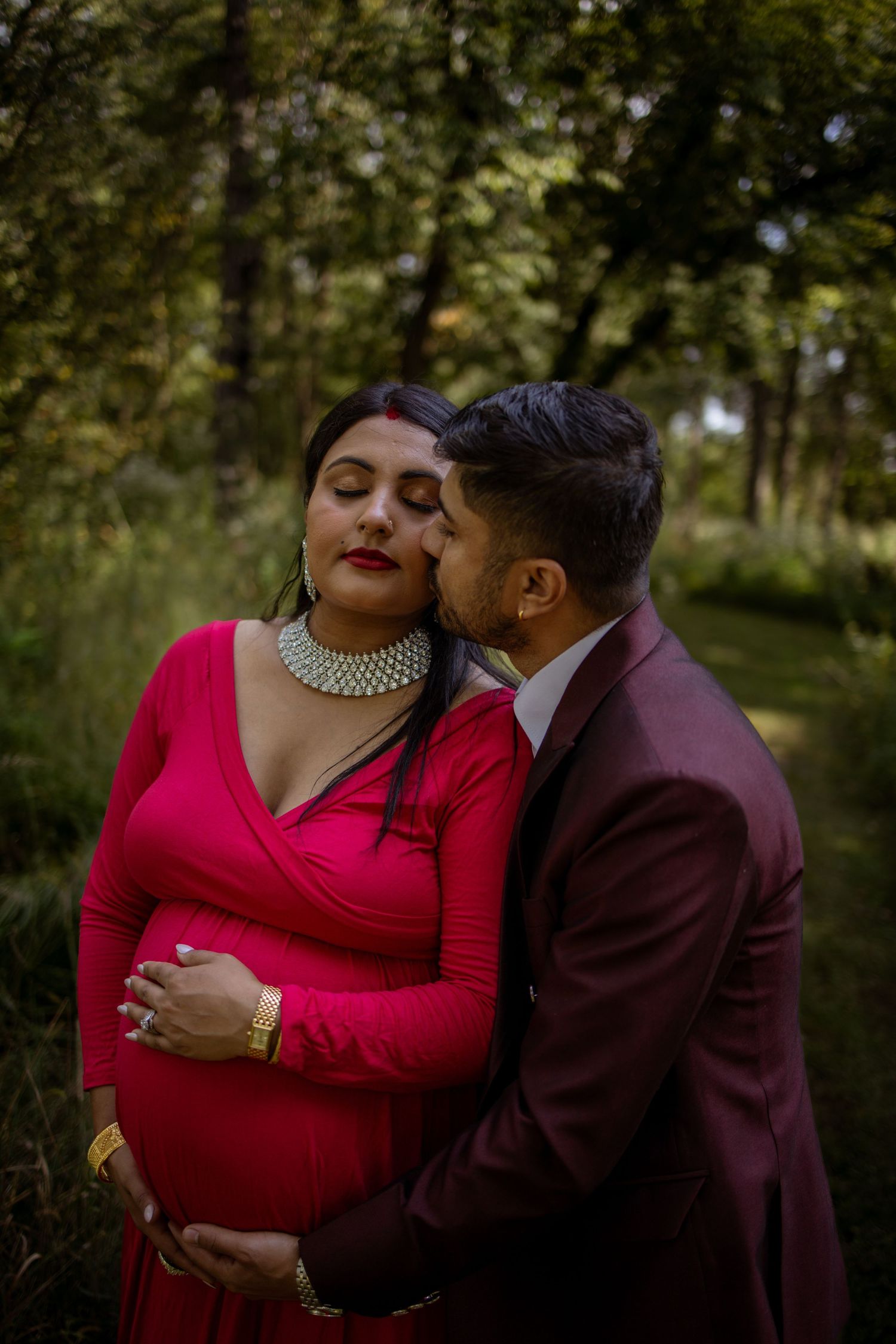 A romantic couple embraces tenderly in a forest setting, with the woman in red and the man in burgundy attire.