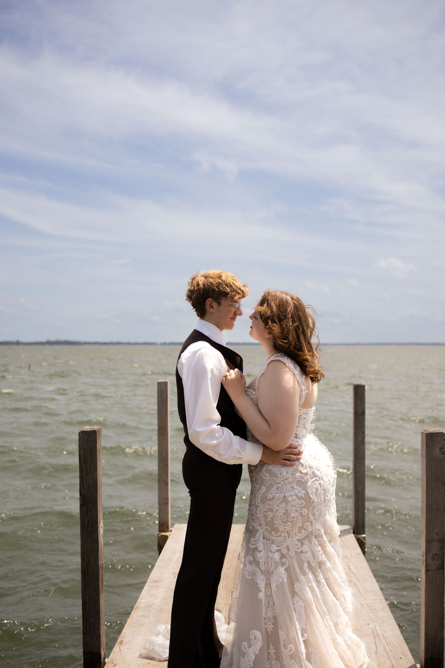 A couple in elegant wedding attire share an intimate moment on a wooden dock overlooking a vast waterfront.