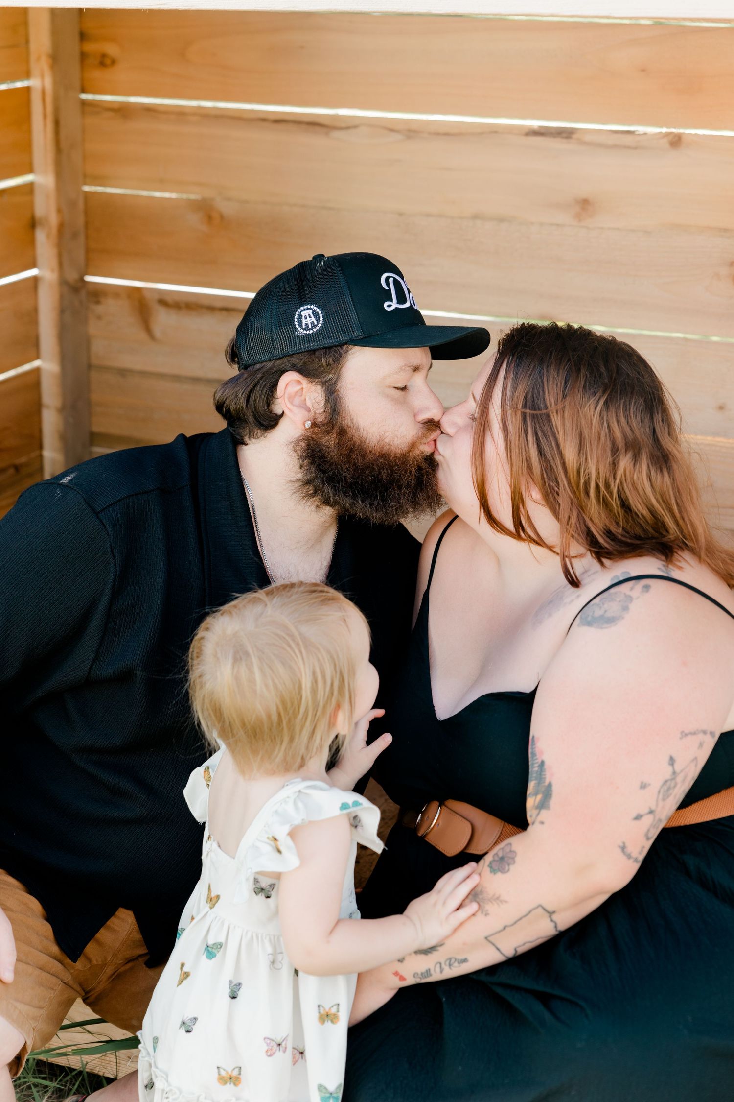 Sweet family moment in wooden interior with infant in white dress and loving parents sharing tender kisses.