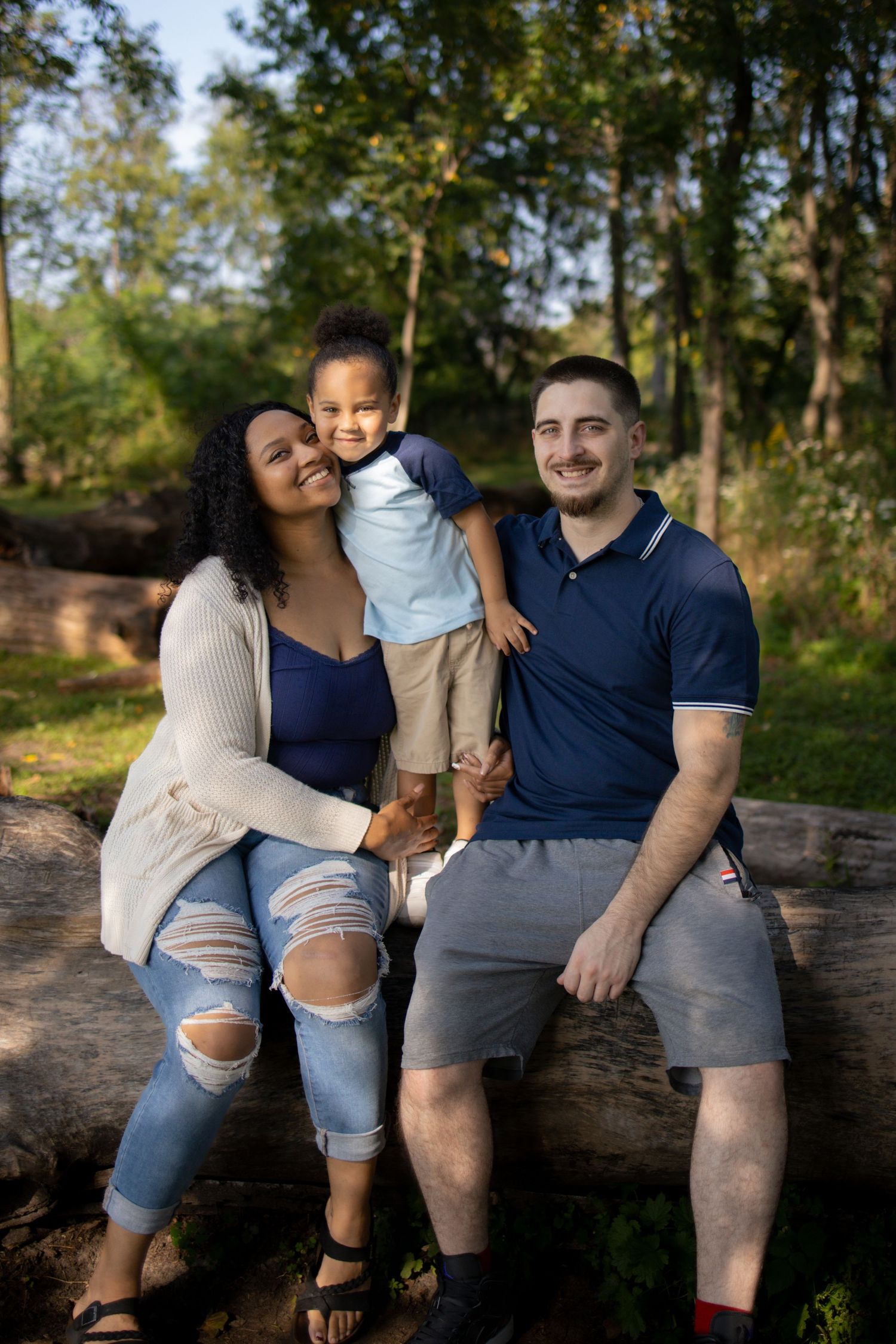 Family sitting together on a rustic wooden log in a sunny wooded area wearing casual denim and navy outfits.