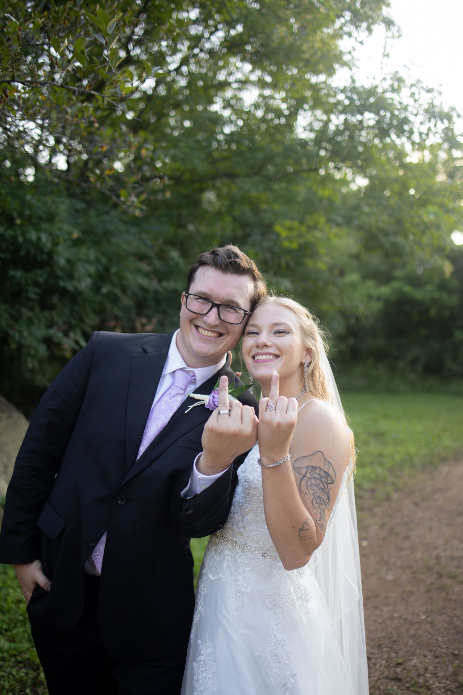 A happy couple showing their wedding rings together on a dirt path surrounded by green trees.