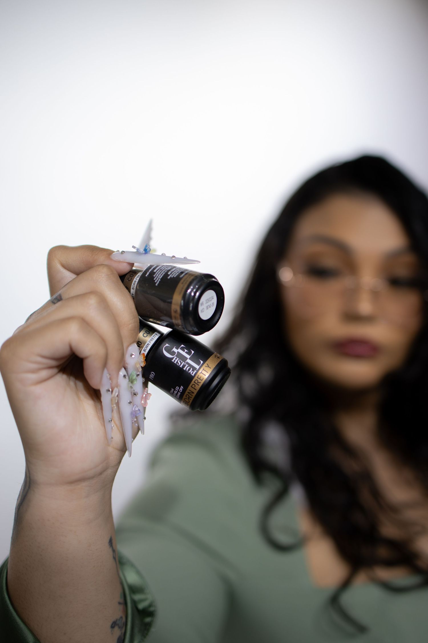 A manicured hand in focus holds up two small vials against a soft blurred background.