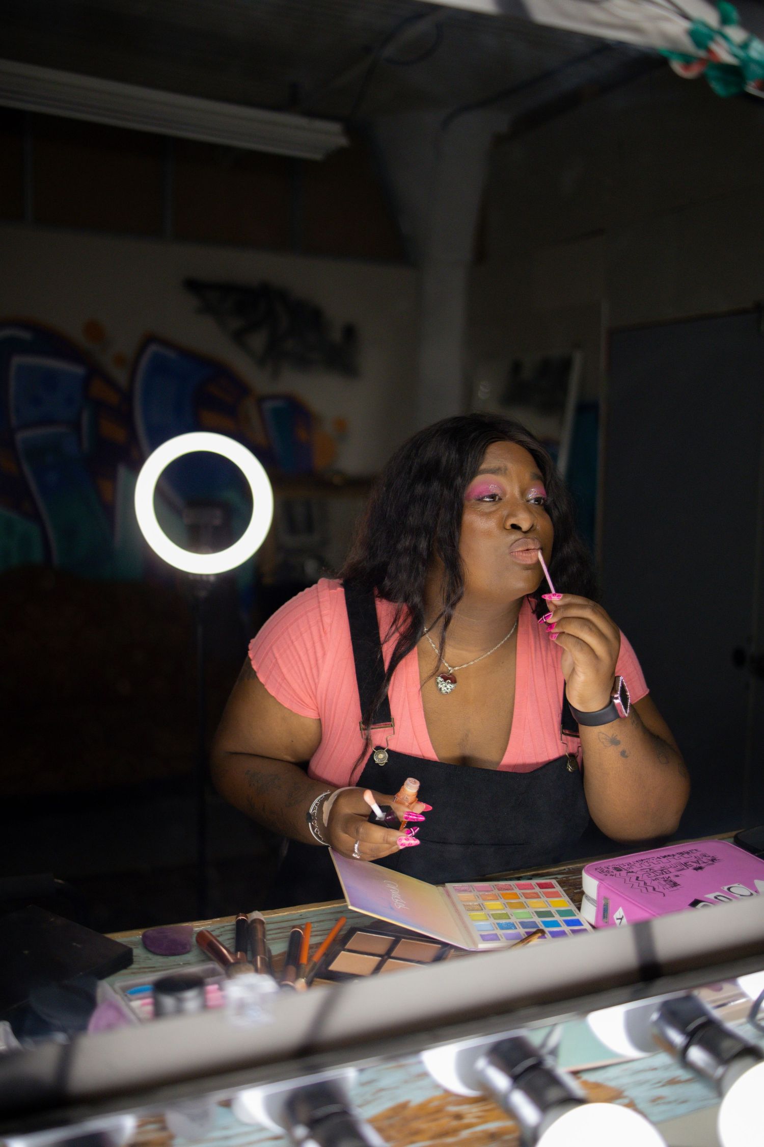 Content creator working at desk with ring light and makeup supplies in dark room with pink notepad.