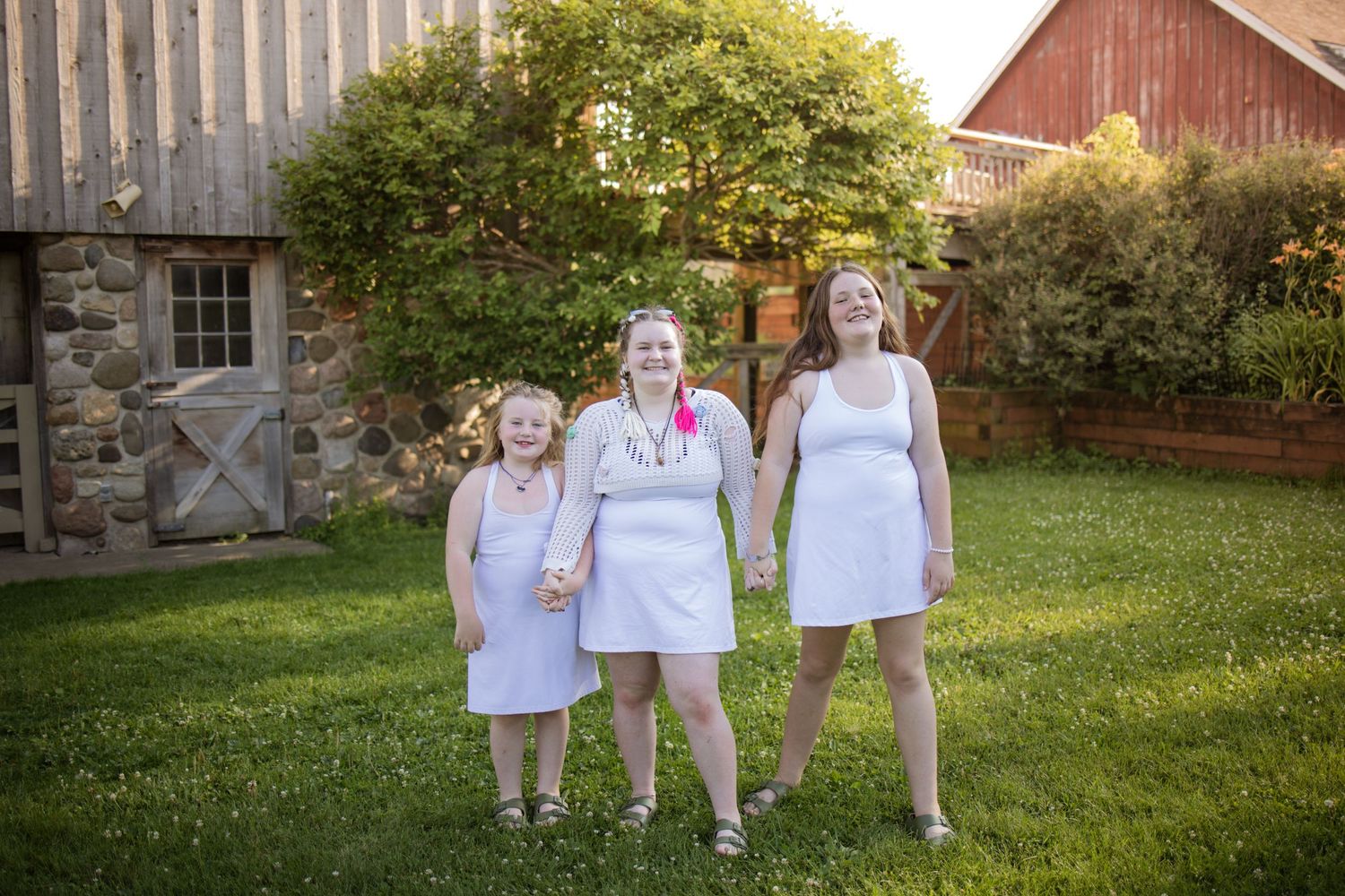 Three people in white dresses stand together on a grassy lawn in front of a rustic stone and wooden barn.