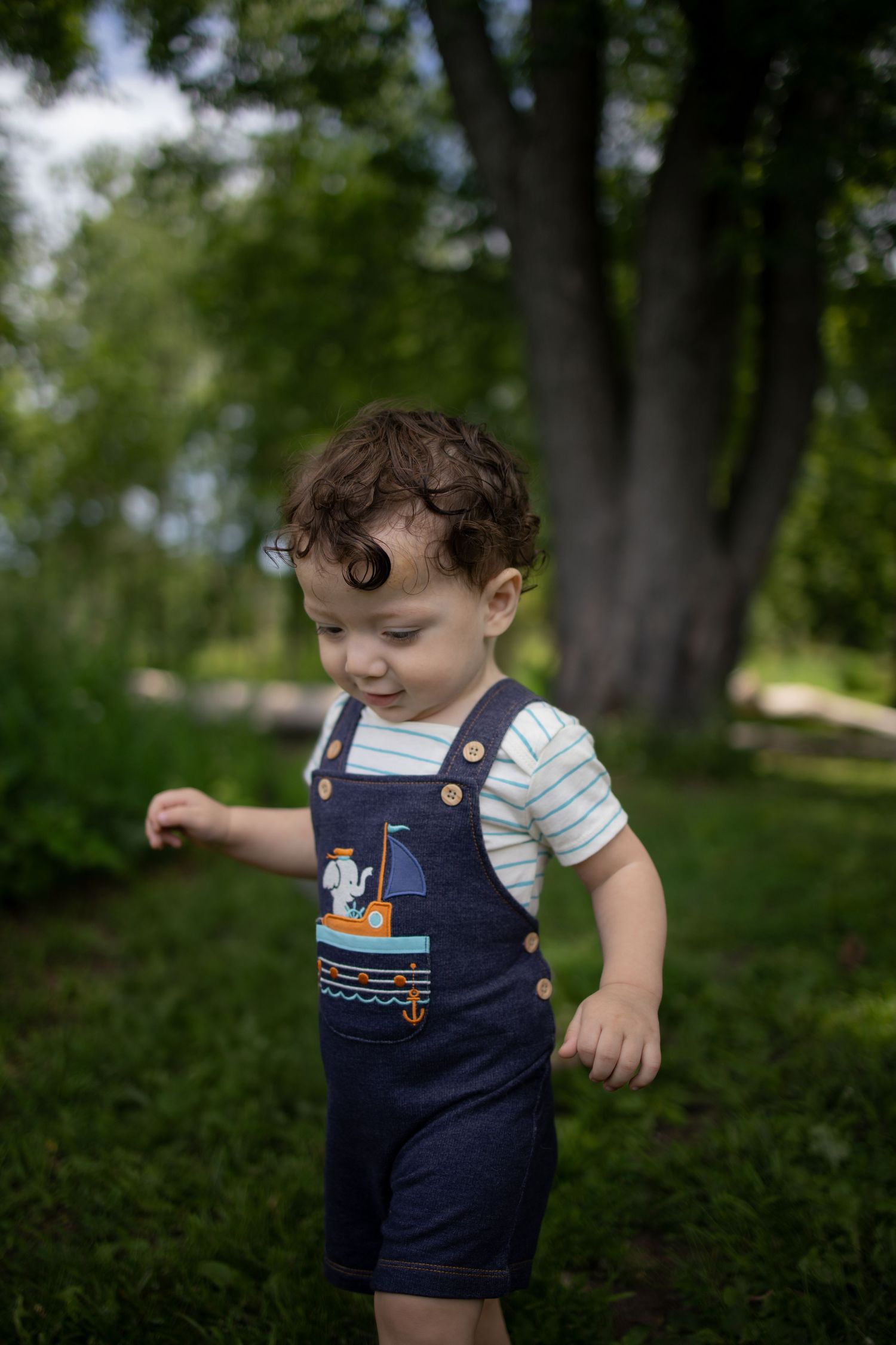 A small toddler in navy blue overalls plays outside in a lush green wooded area on a sunny day.