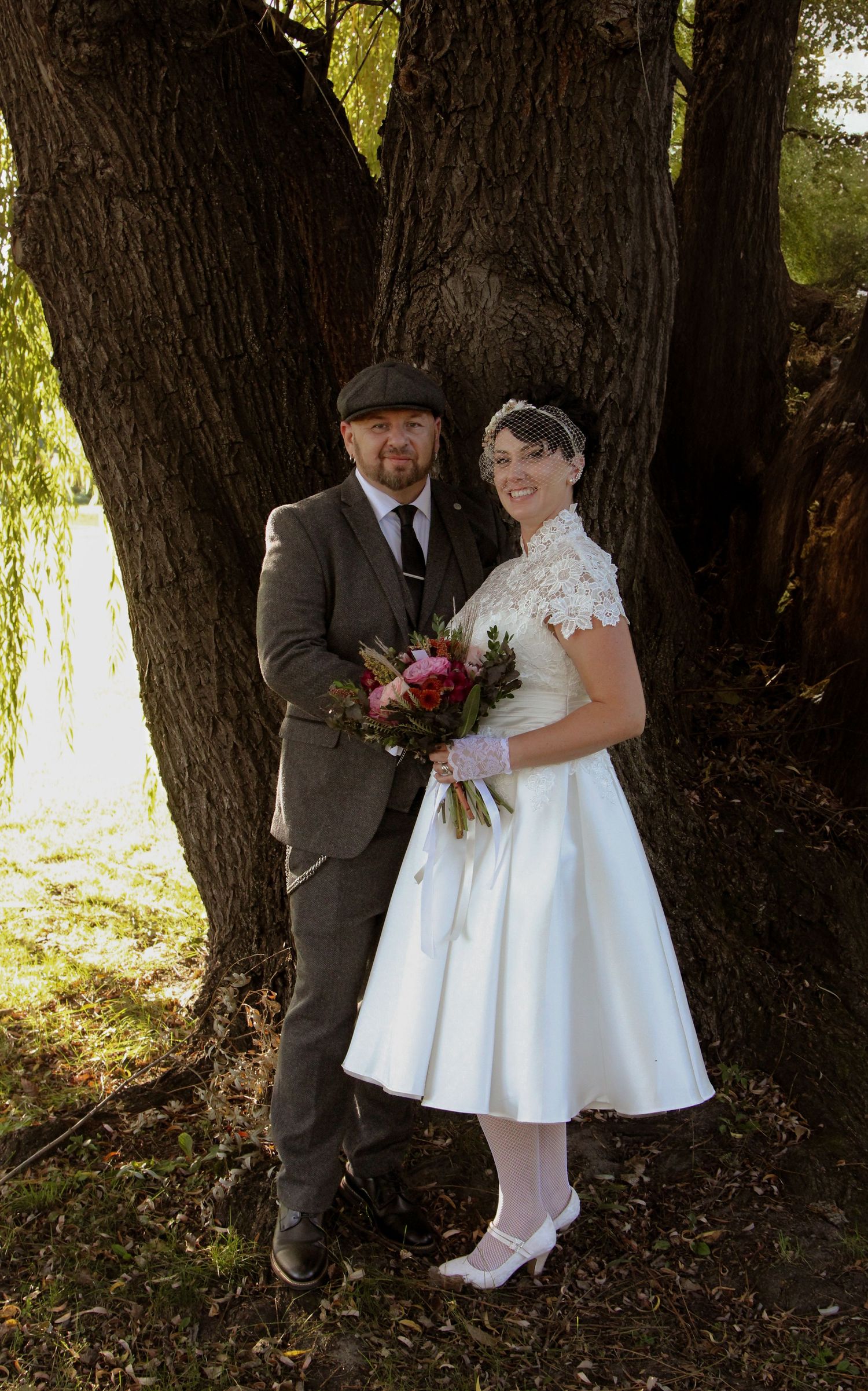 A couple poses together by a large tree trunk in vintage-inspired wedding attire including a tea-length dress and tweed suit.