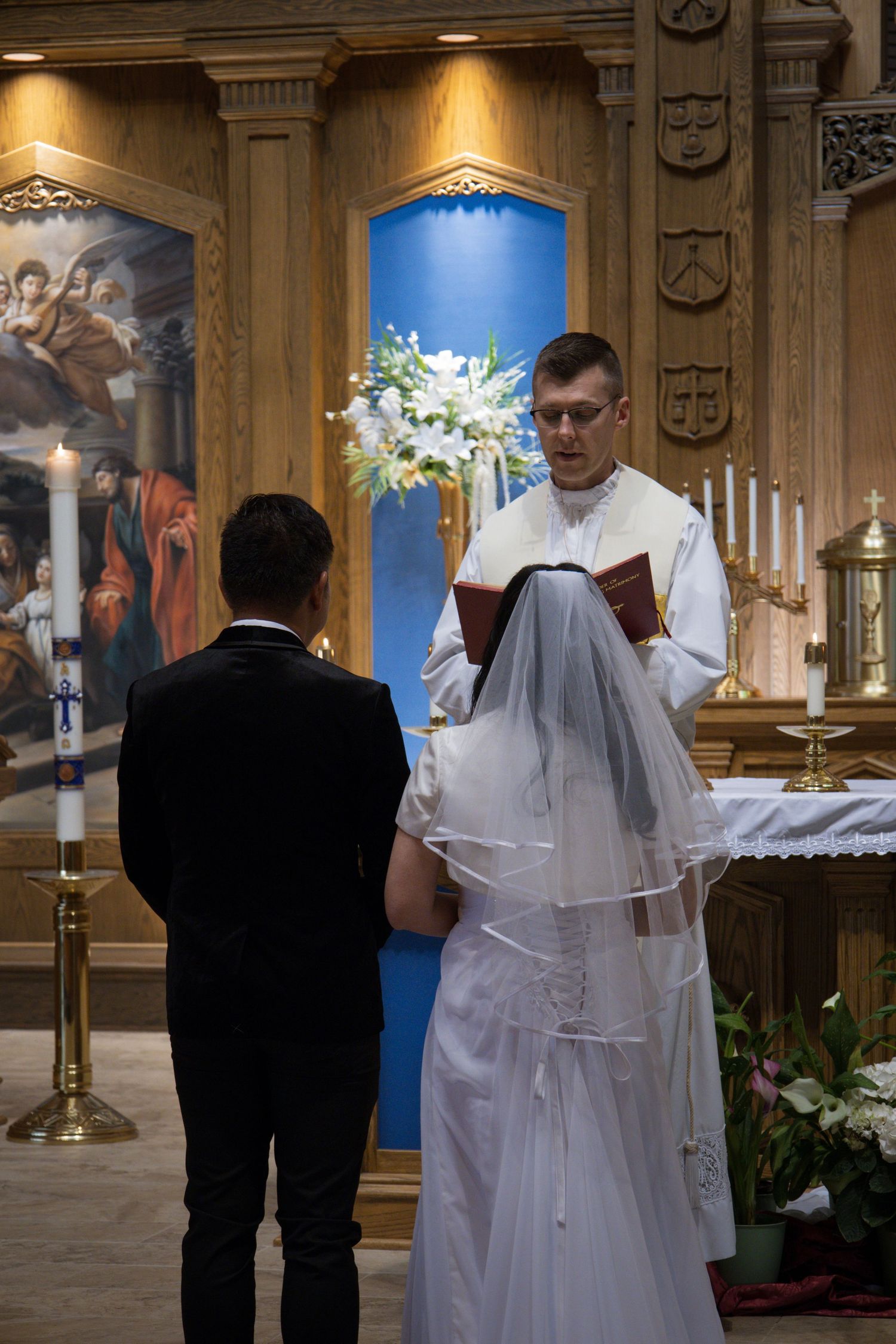 A wedding ceremony takes place inside an ornate church with a floral arrangement and candles on the altar.