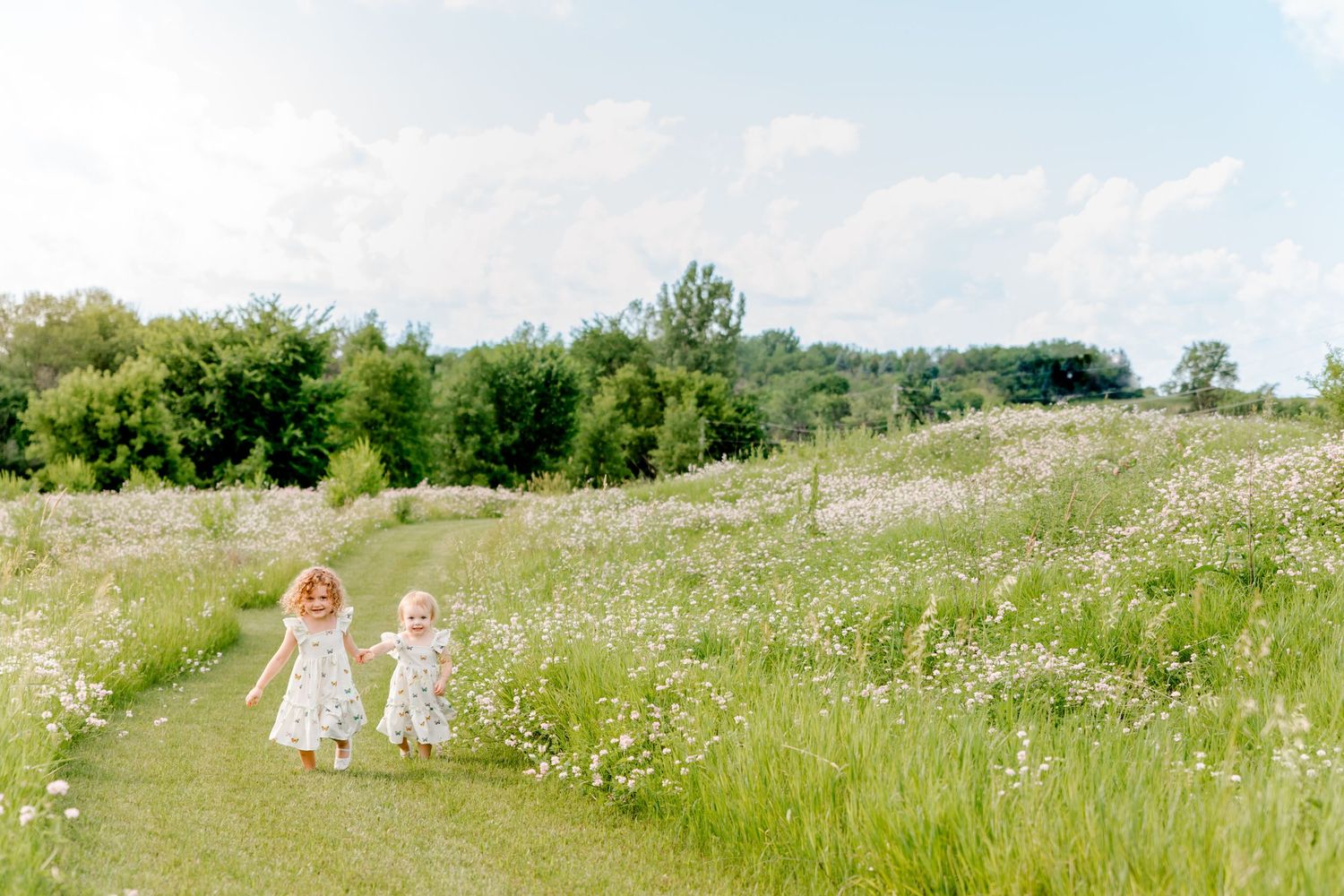 Two children in white dresses walk through a sunny wildflower meadow on a summer day.