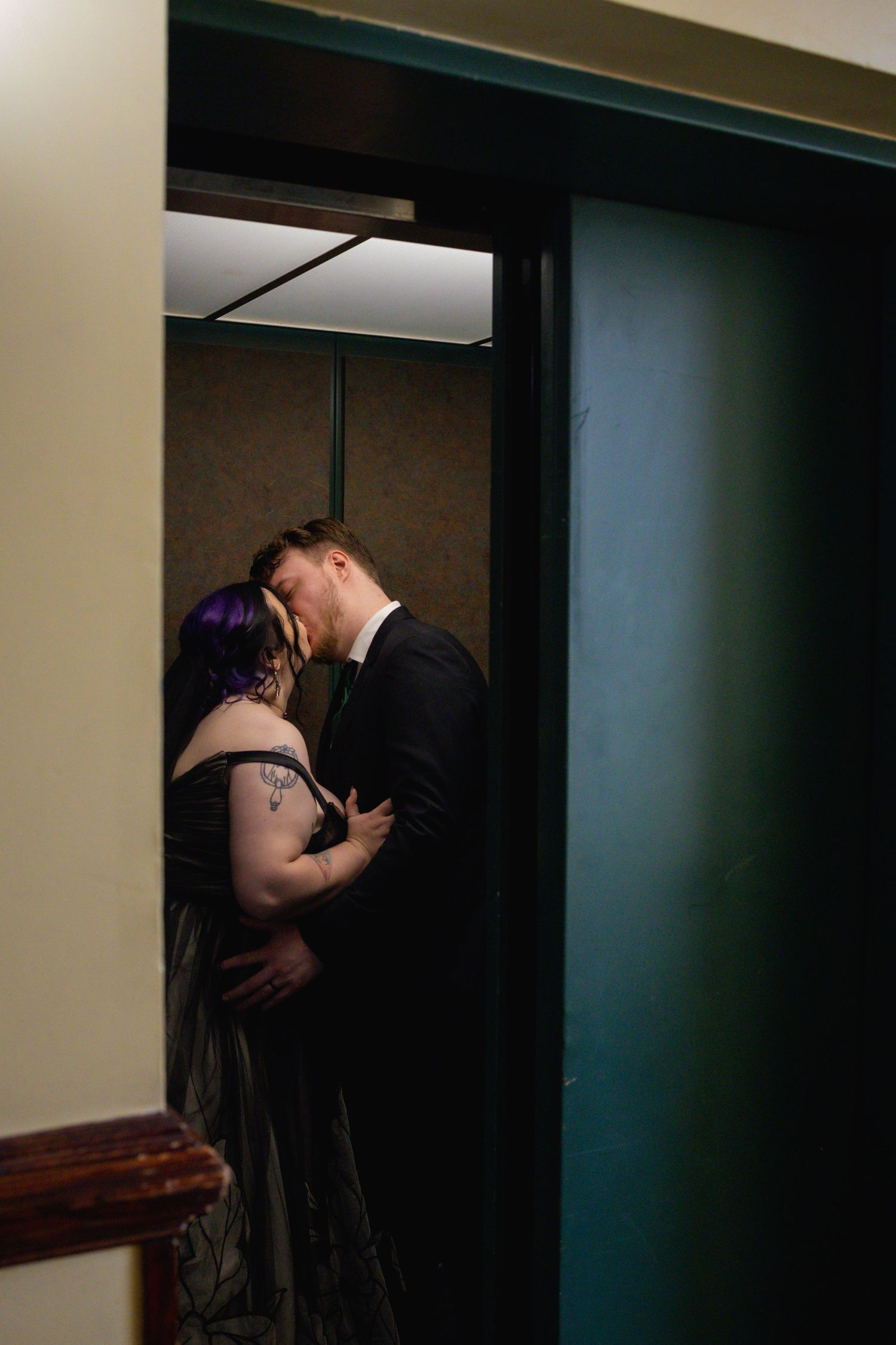 A romantic couple shares an intimate moment in a dimly lit doorway, with one person wearing purple hair and a dark dress.