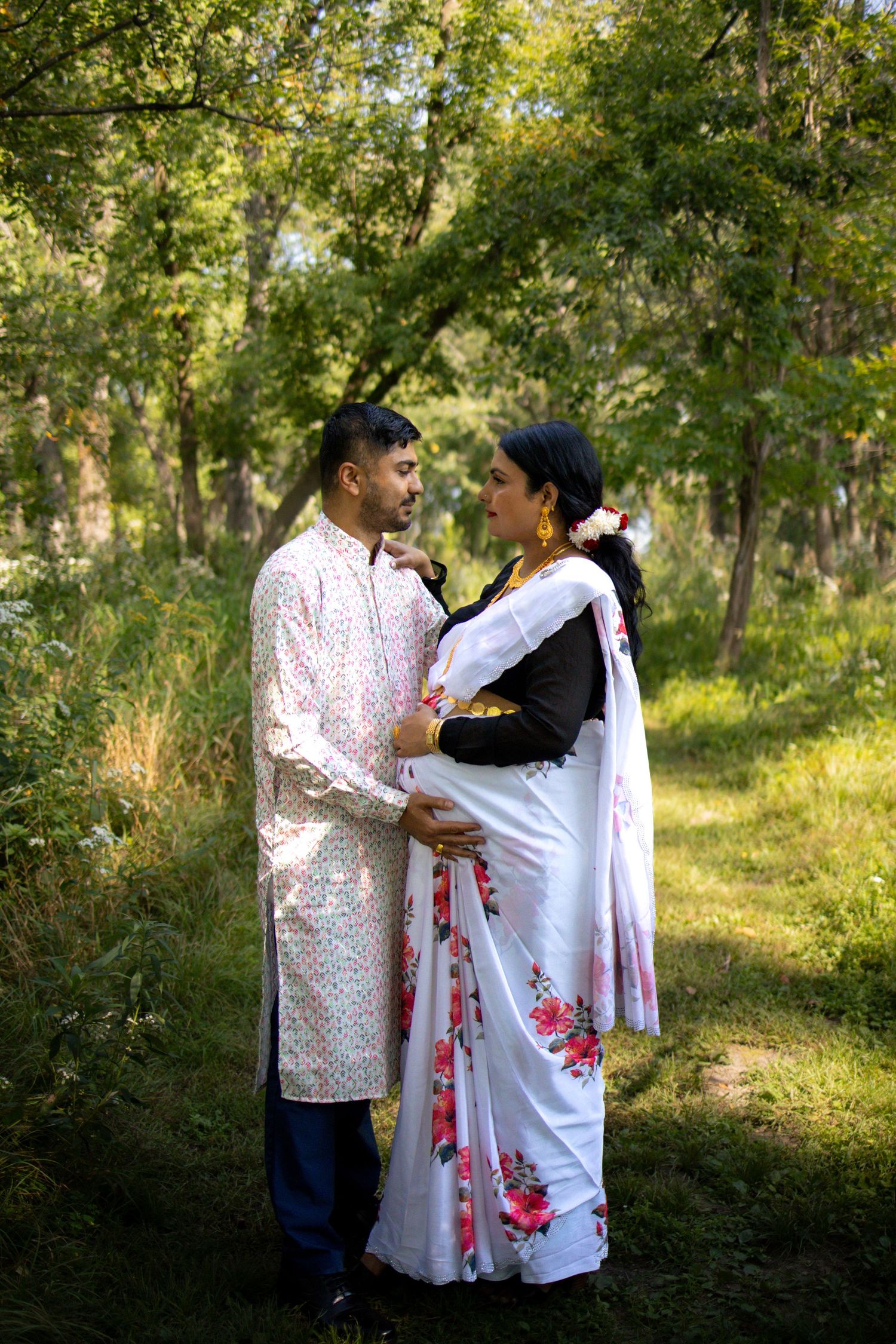 A couple shares an intimate moment in traditional white outfits with red floral details amid a wooded outdoor setting.