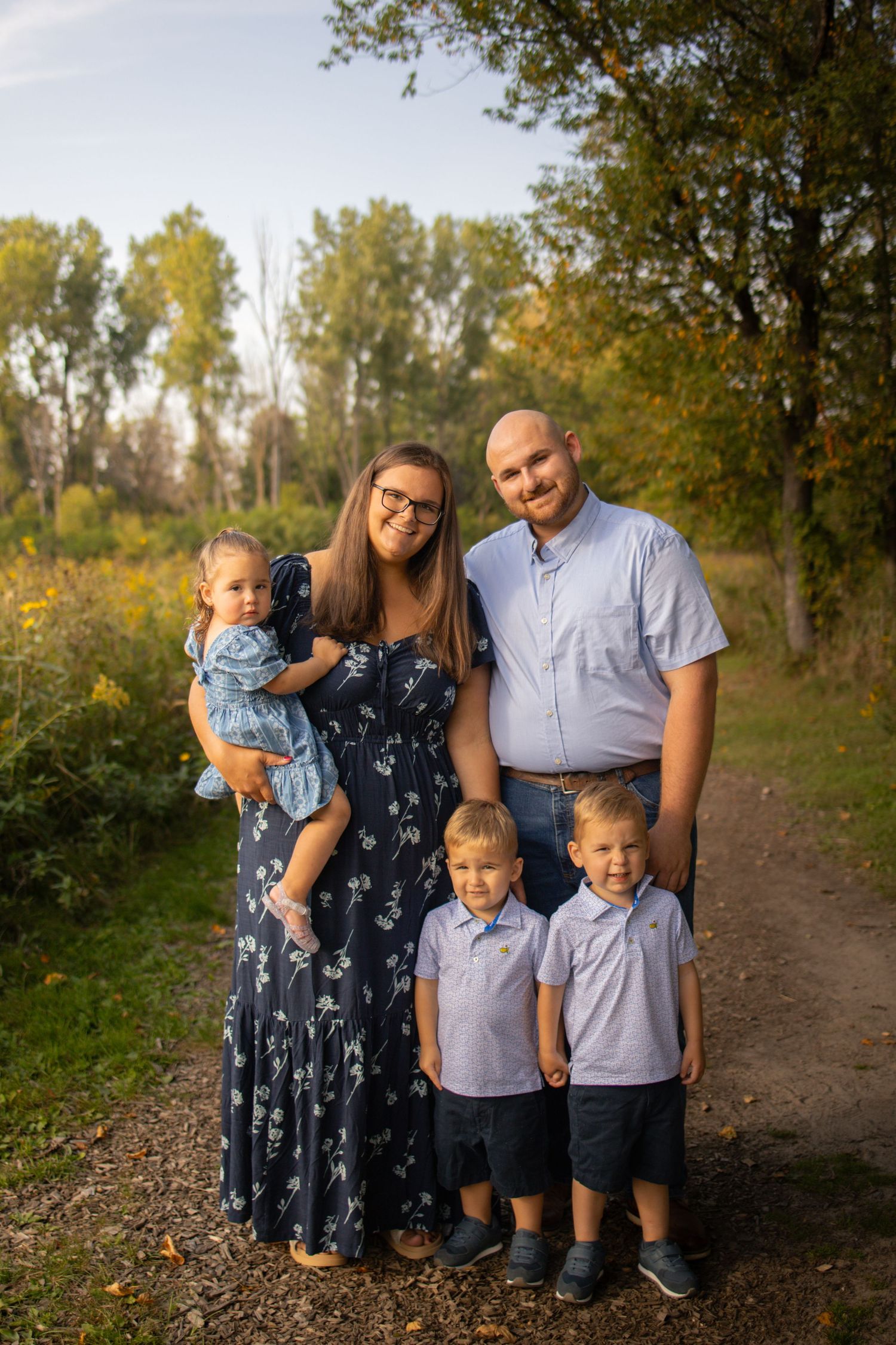 A family wearing coordinated navy blue and light blue outfits poses together on a wooded path during autumn.