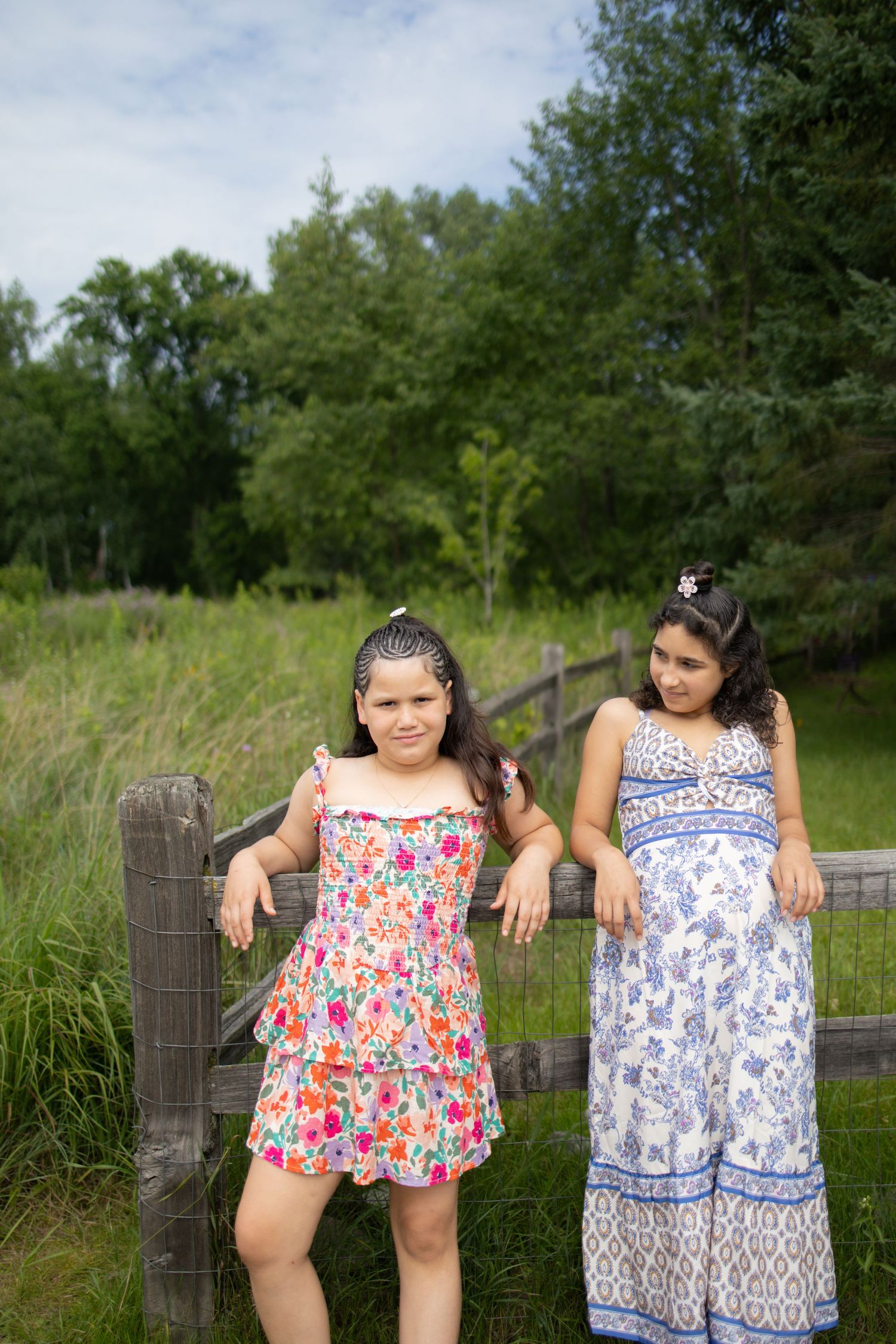 Two people in floral print sundresses lean against a wooden fence post in a grassy summer field.