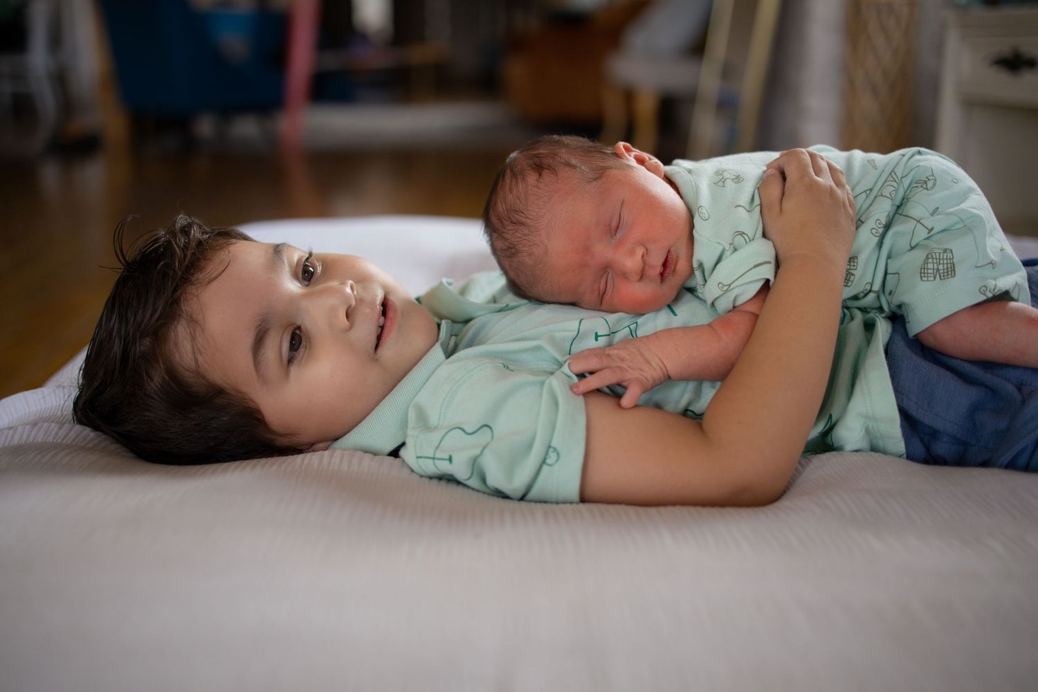 A young child lies on a bed holding a sleeping newborn baby wearing teal hospital clothing.
