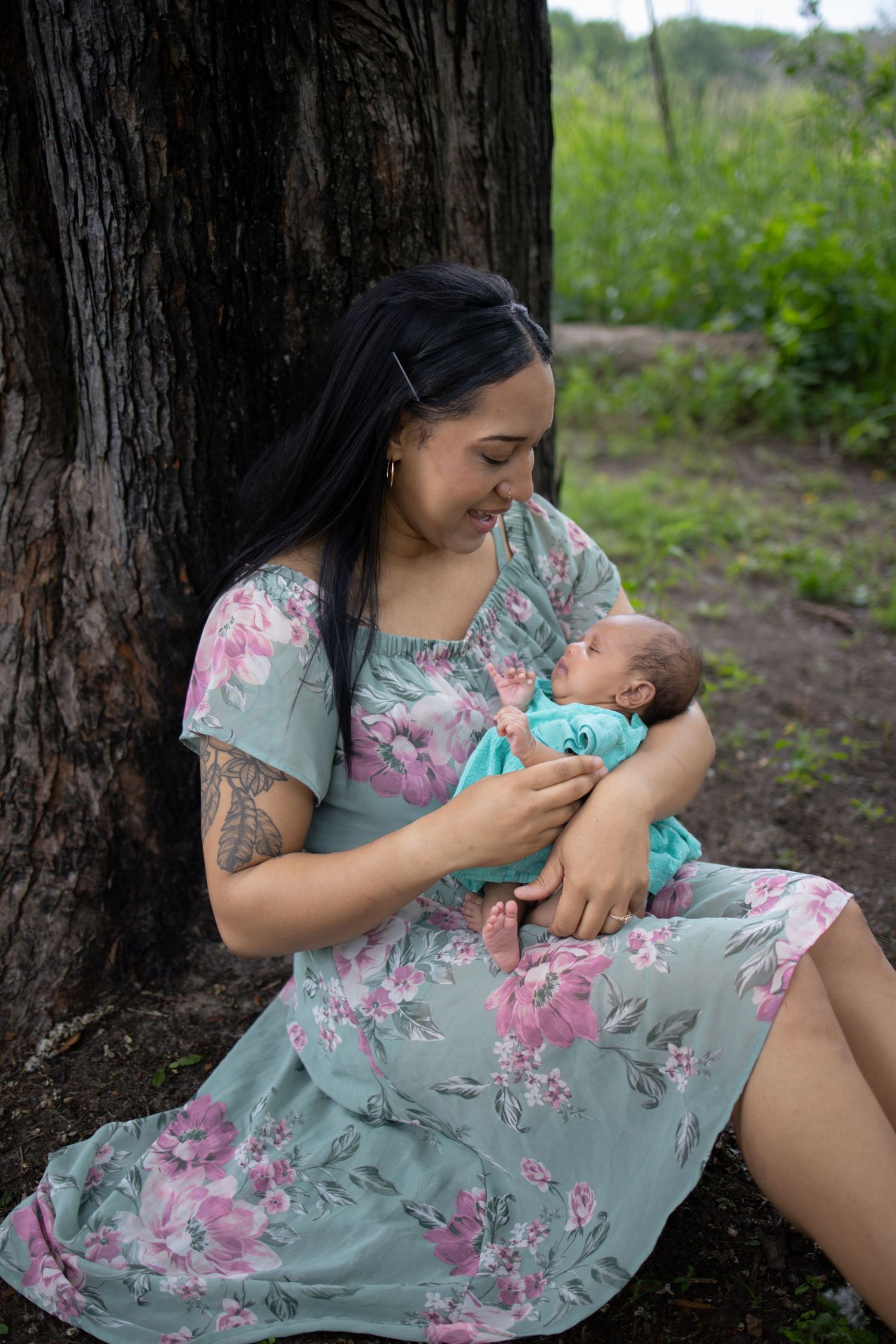 A person wearing a floral mint green dress sits against a tree trunk while feeding an infant outdoors.