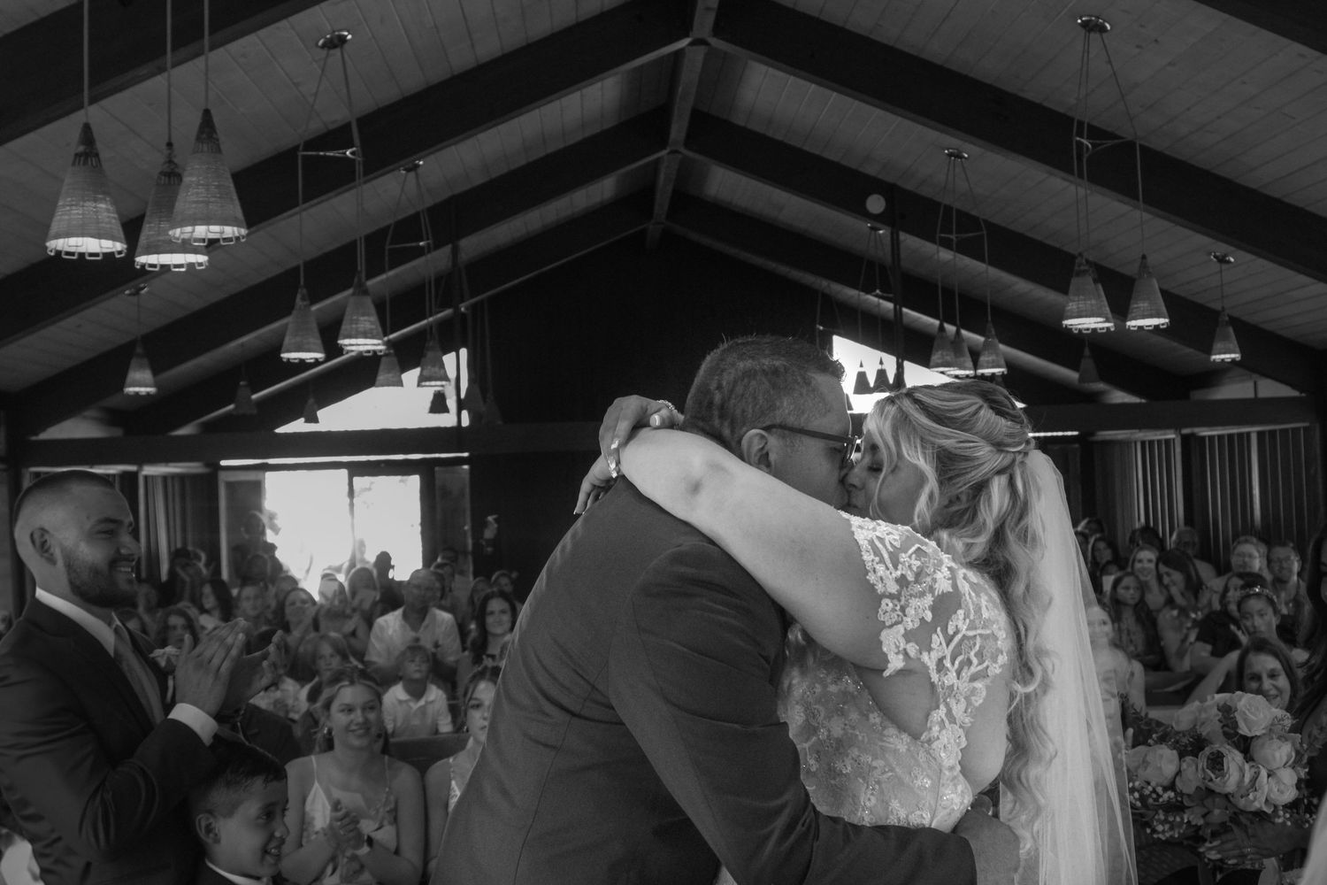 Large church ceremony with guests standing together under wooden beams and pendant lights during a celebration.