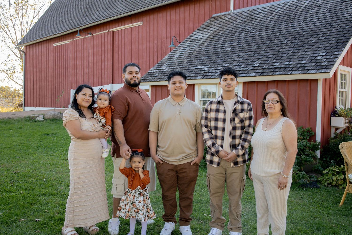 A group of people in casual attire pose together on a grassy lawn in front of a red barn on a sunny day.