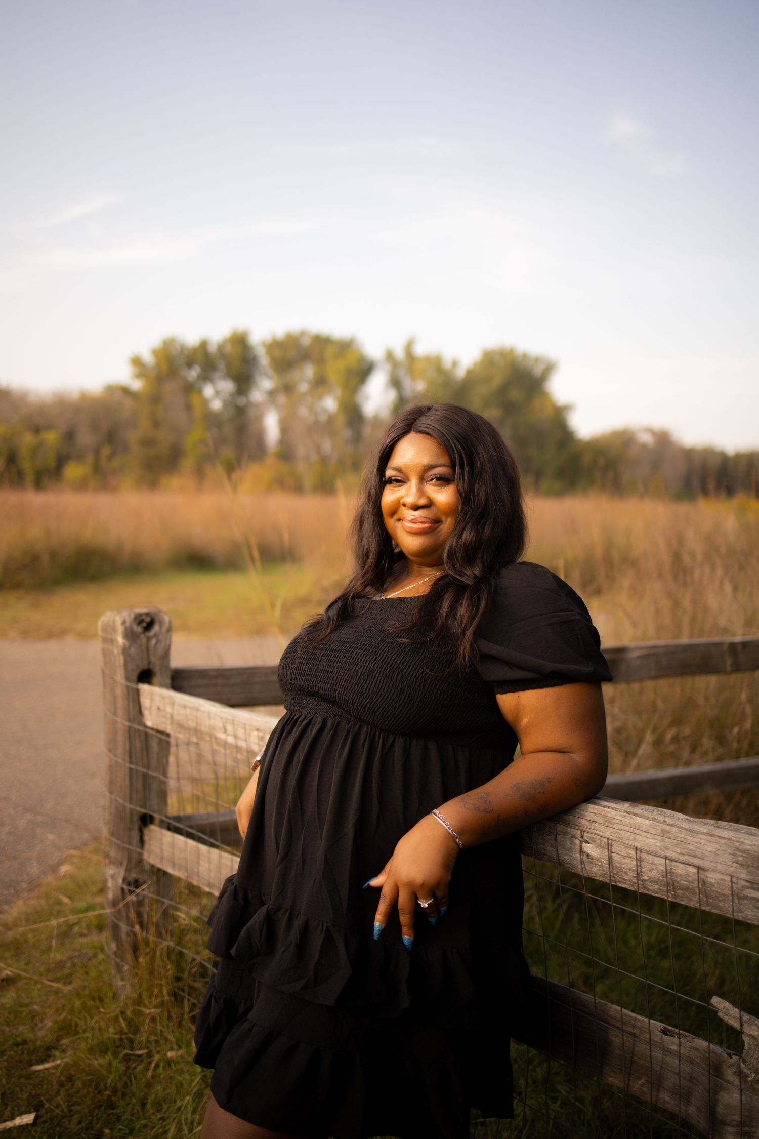 Person in a flowing black dress poses outdoors against a rustic wooden fence during golden hour.