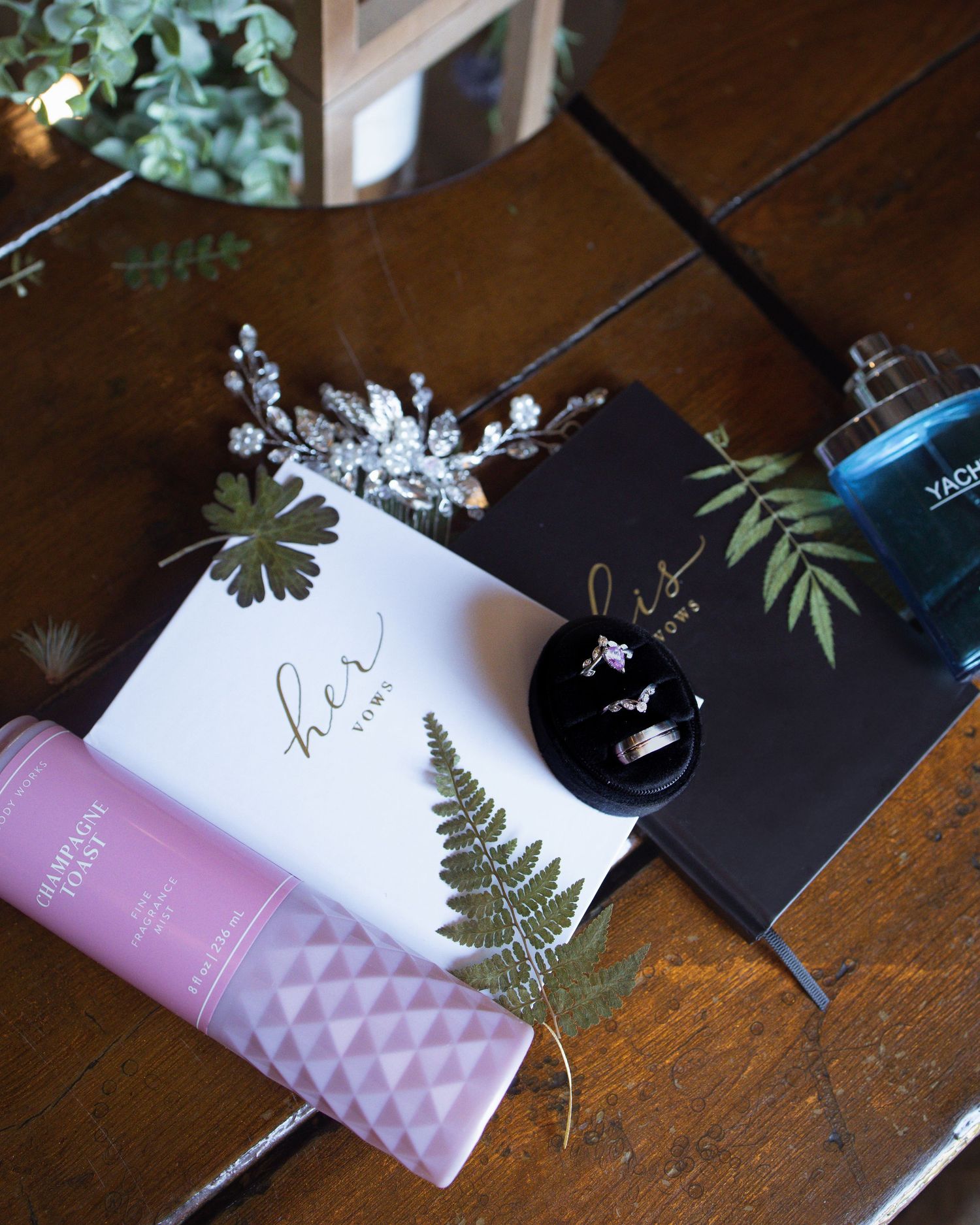 Wedding accessories including jewelry, fern leaves, and a pink bottle arranged on a dark wooden surface.