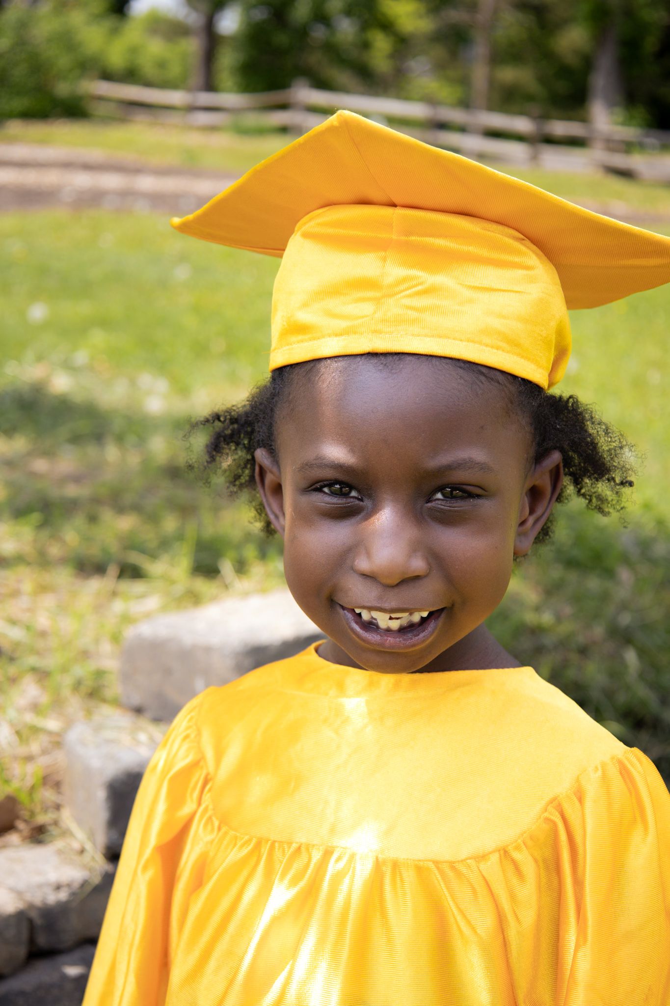 Young graduate wearing bright yellow cap and gown smiles warmly outside on a sunny day.
