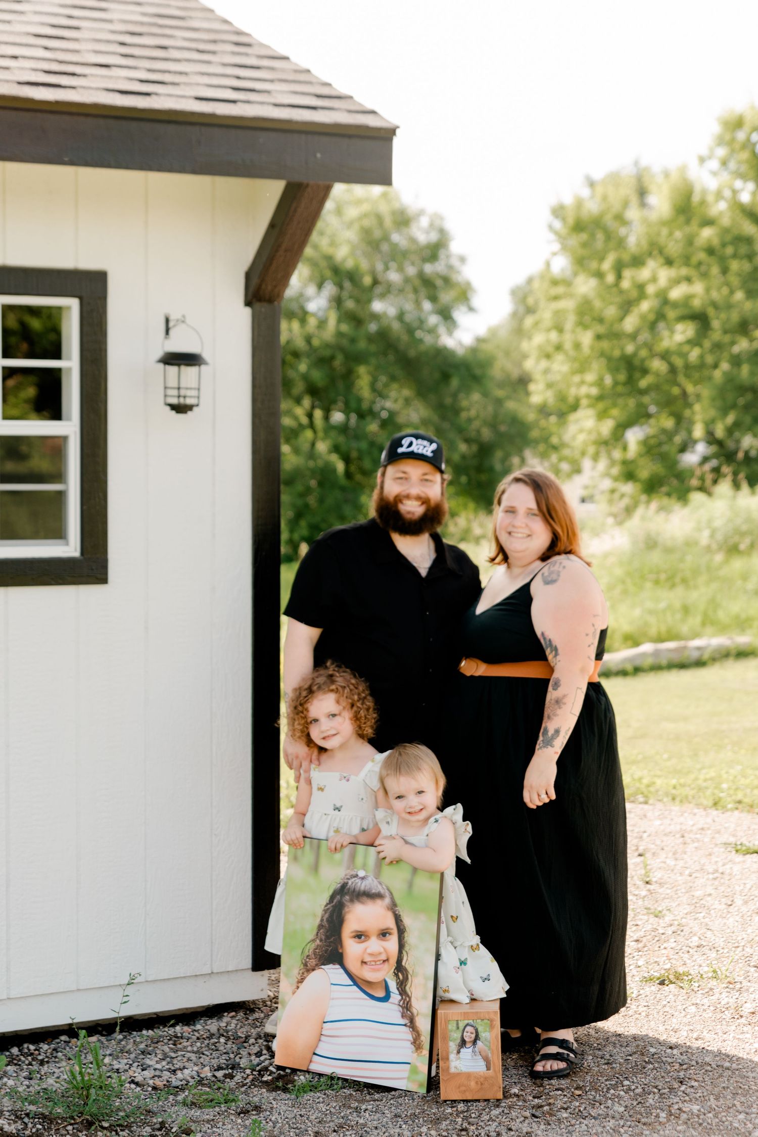 A family poses together beside a white house with black trim and a wall-mounted lantern on a sunny day.