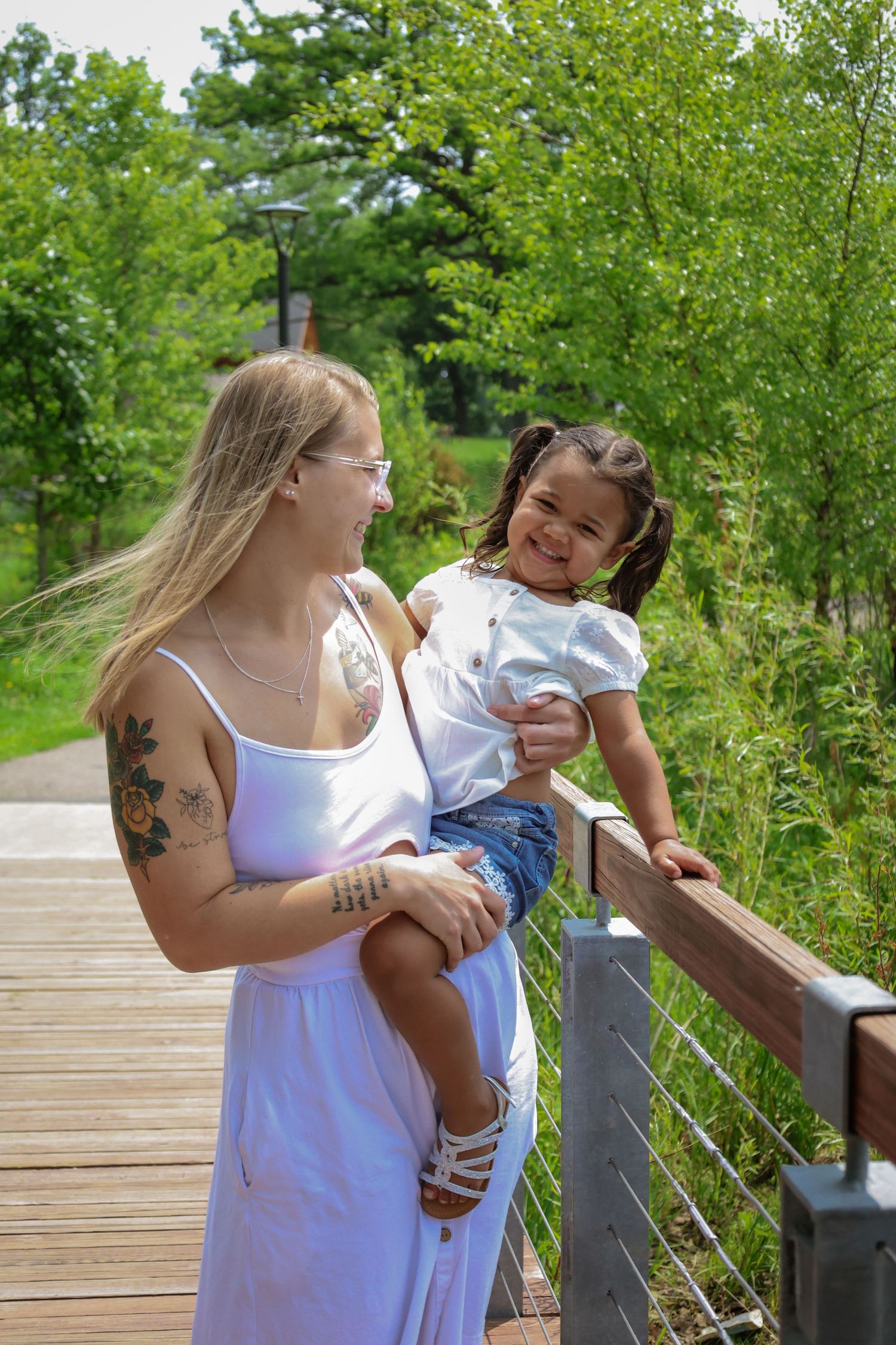 A woman in a white dress shares playful moments with a small child on a wooden boardwalk in a lush green setting.