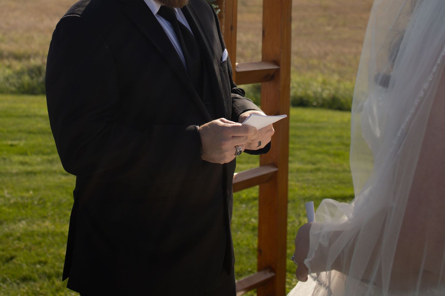 A couple in formal wedding attire stands together on a porch with sunlit grass in the background.