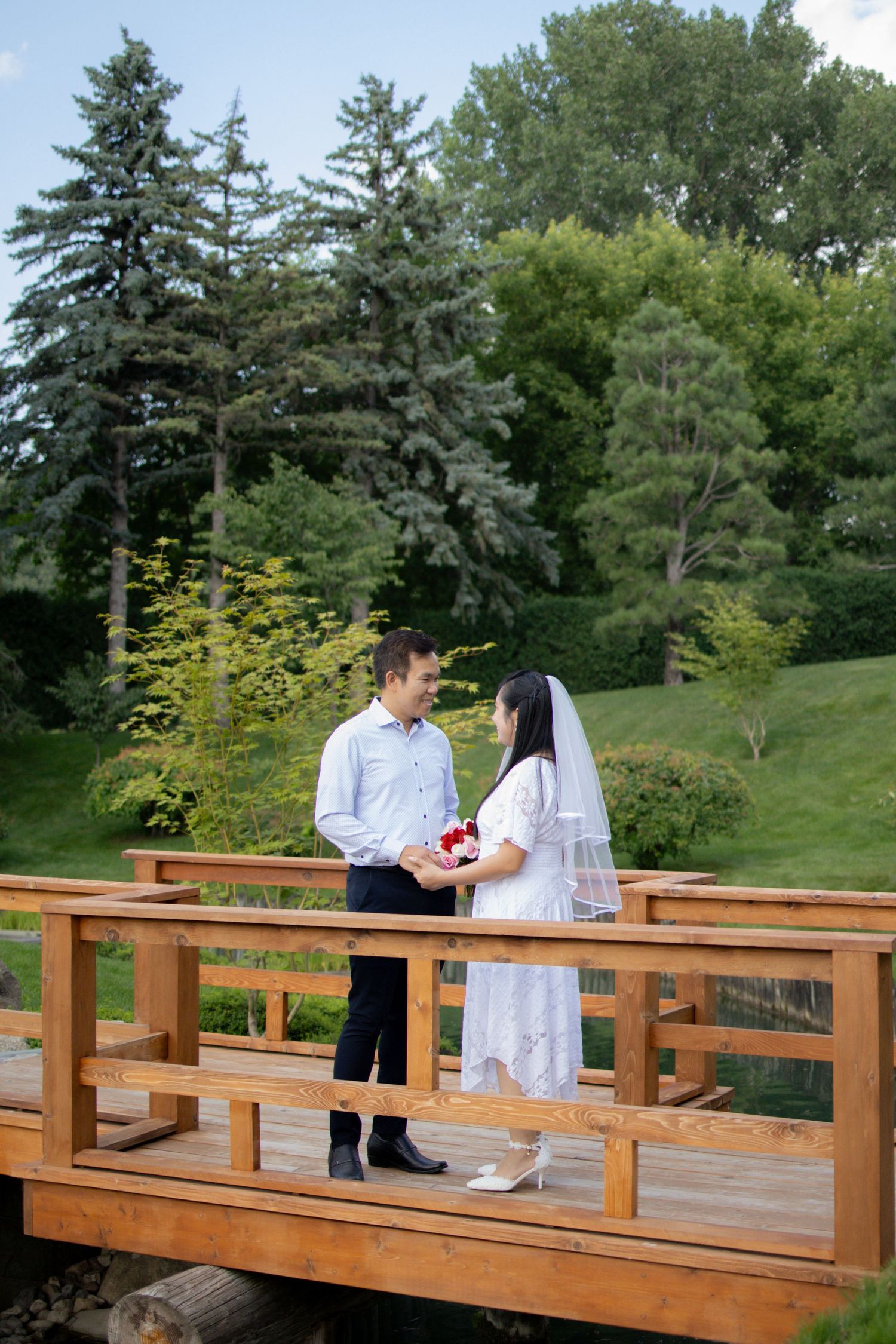 A couple shares a romantic moment on a wooden bridge surrounded by evergreen trees and lush greenery.