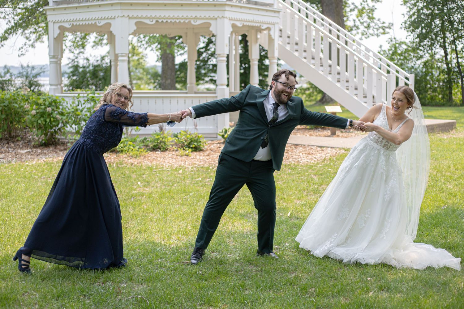 Three people in formal attire pose playfully outdoors near white gazebo stairs during wedding celebration.