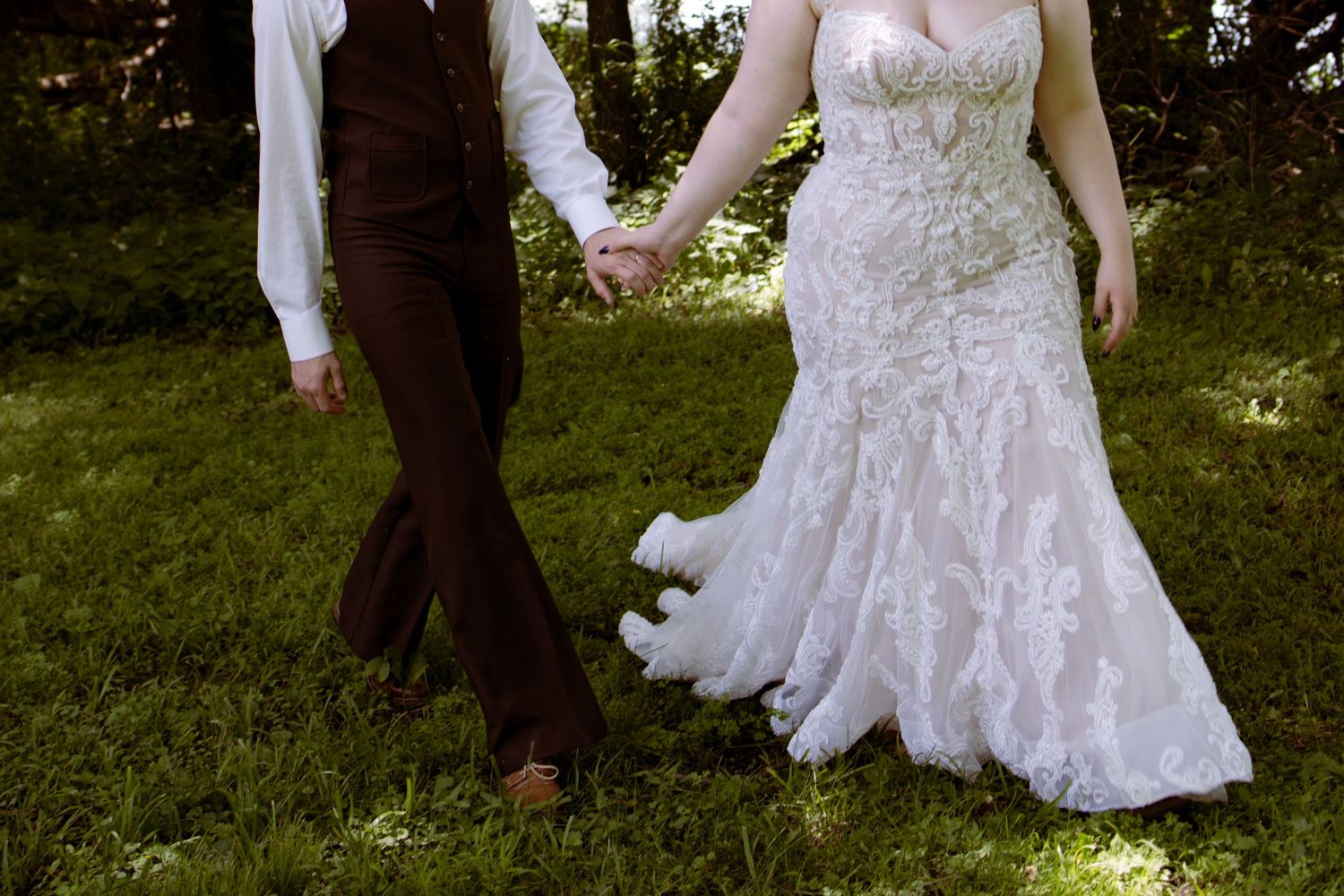 A close-up of a couple holding hands outdoors, showing a brown suit and white lace wedding dress.
