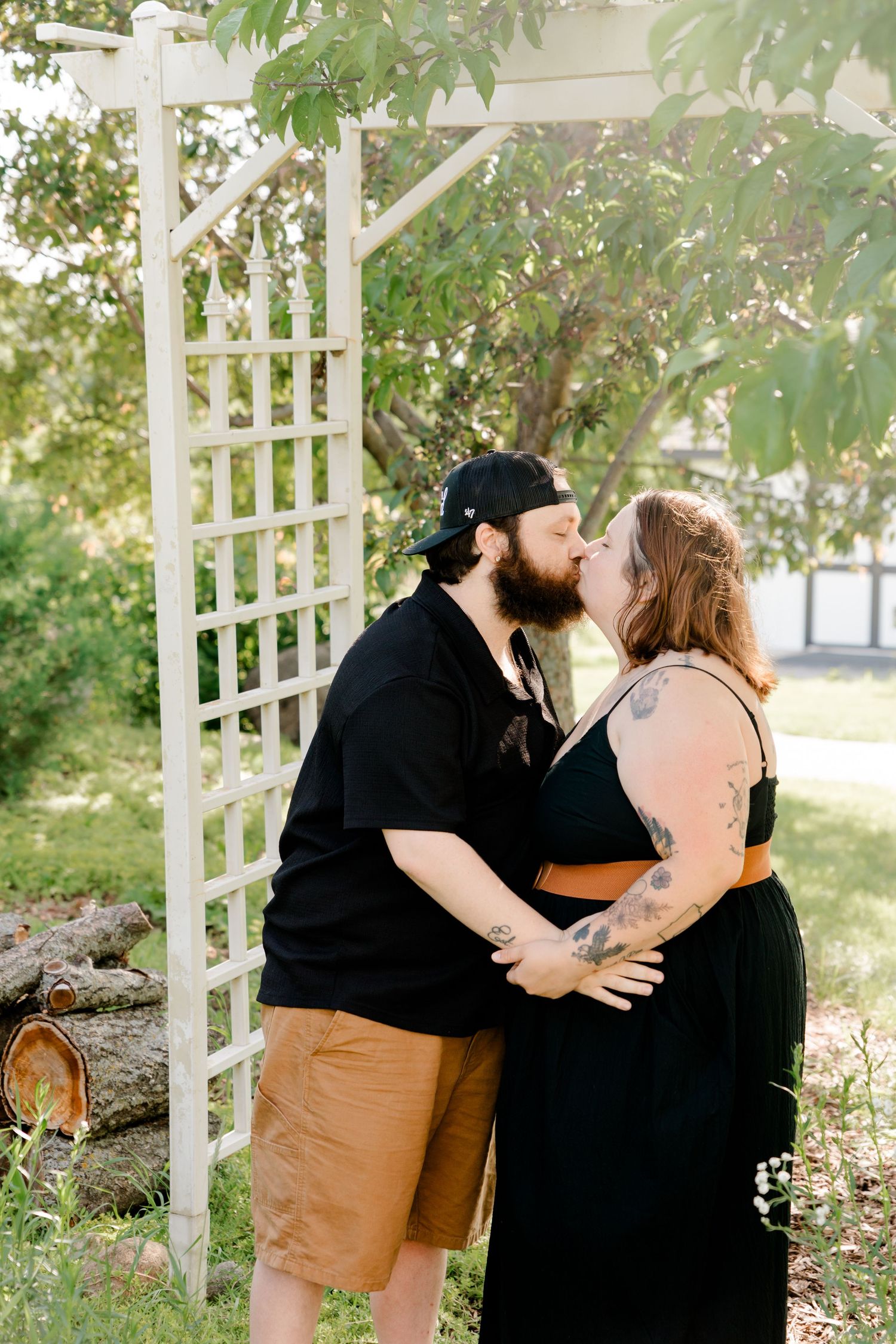 A couple shares an intimate moment under a white garden trellis on a bright summer day.