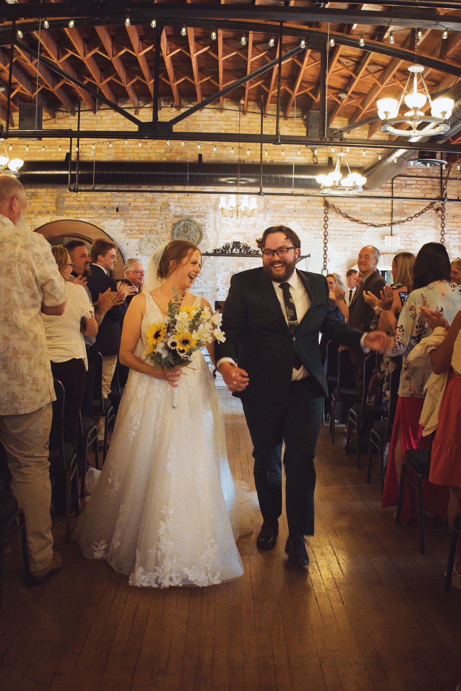 A bride in a white lace dress with yellow bouquet walks down the aisle in a rustic brick venue with exposed wooden beams.