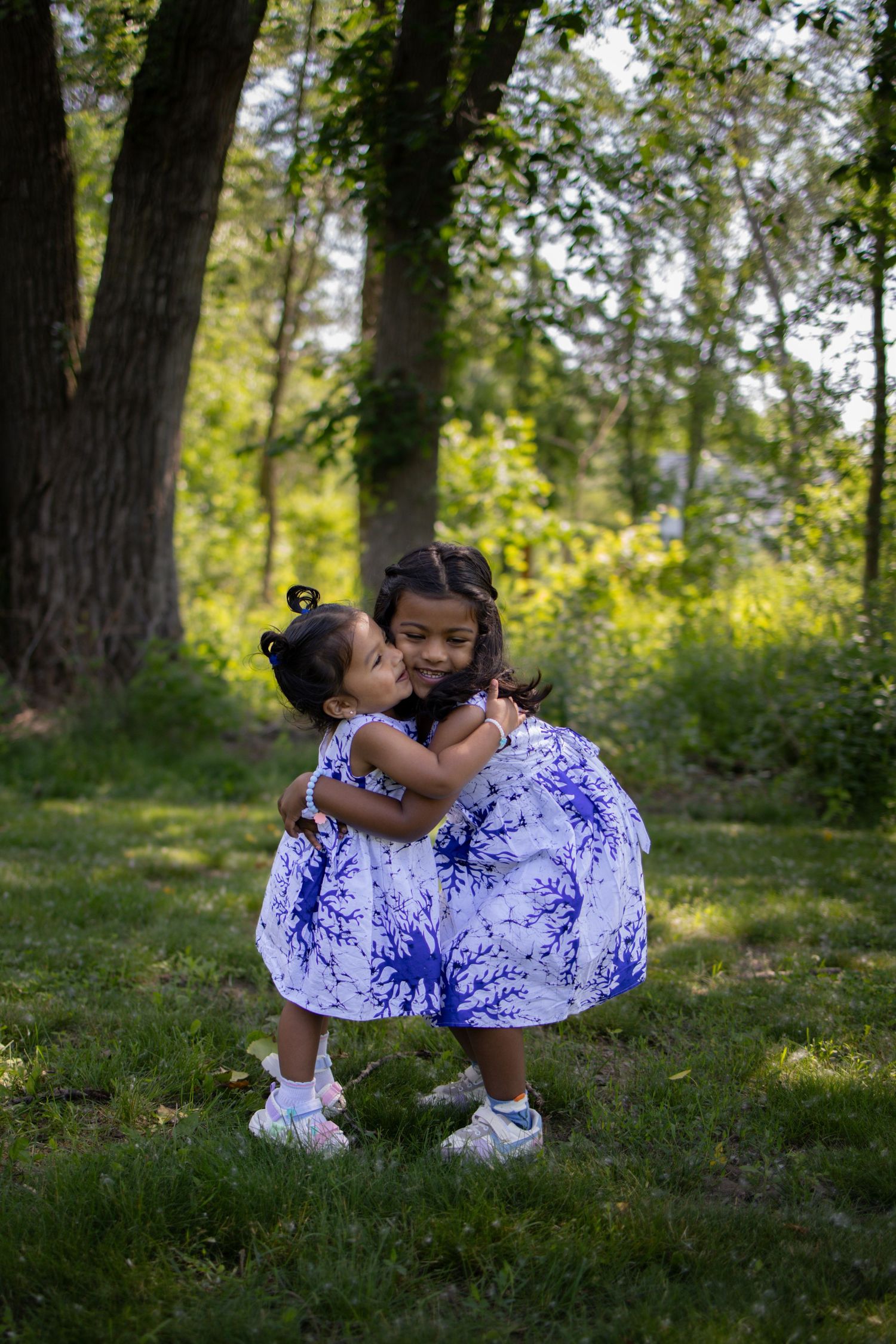 Two children sharing a sweet hug outdoors while wearing matching blue and white dresses in a wooded park setting.