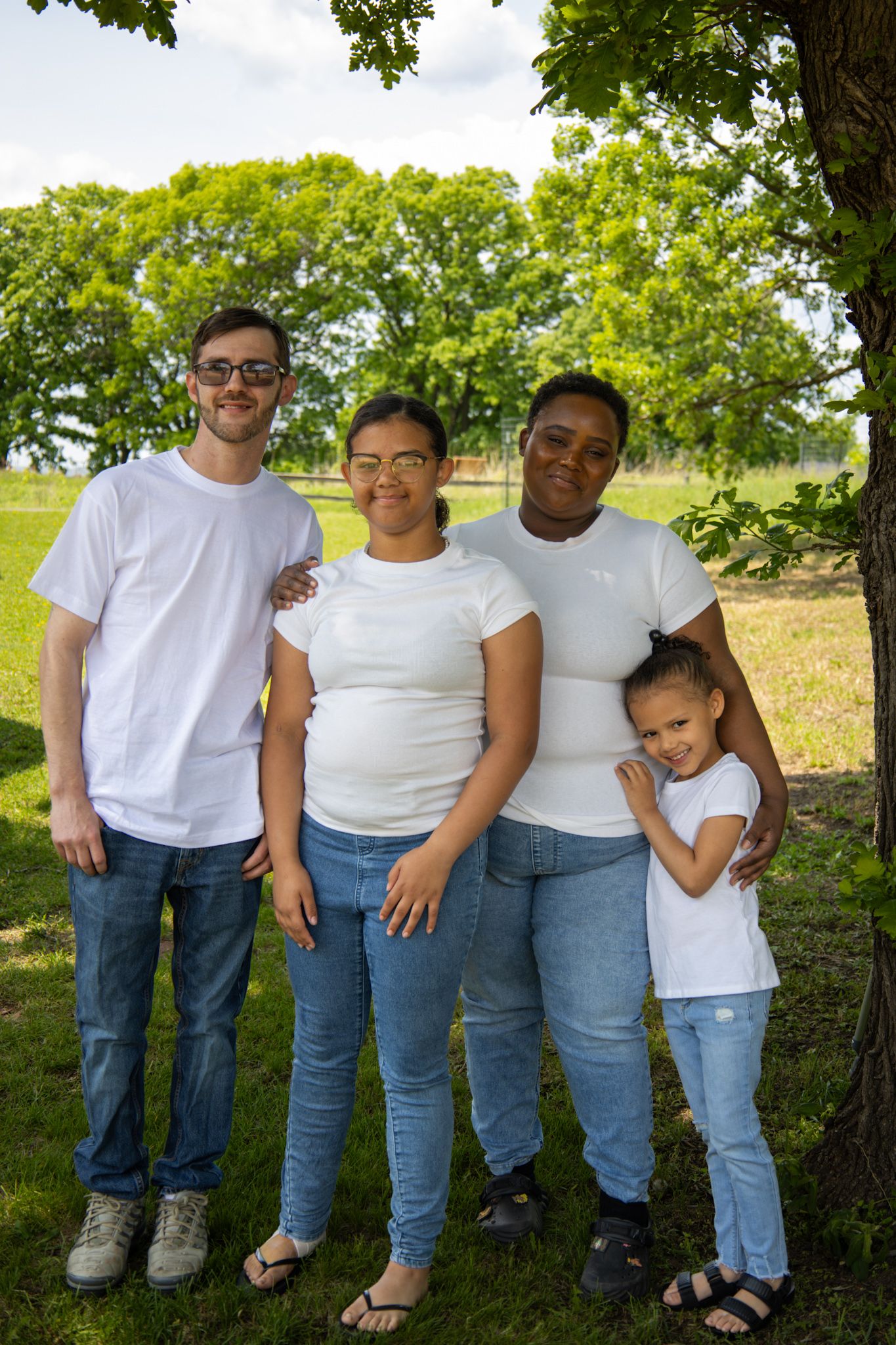A group poses together outdoors in white t-shirts and jeans against a backdrop of green trees on a sunny day.