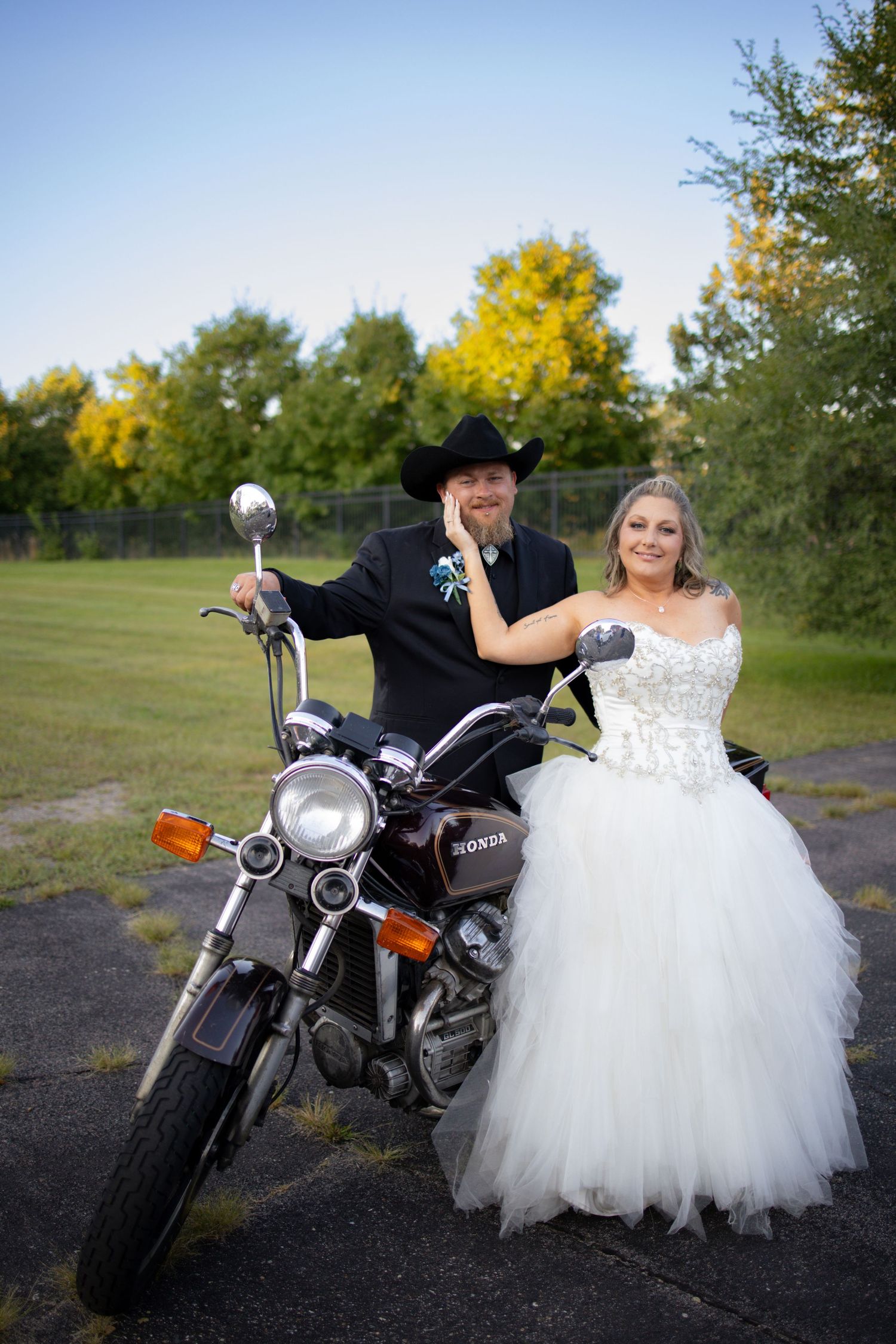 Wedding couple poses creatively with black motorcycle in outdoor setting with ballgown dress and leather jacket.