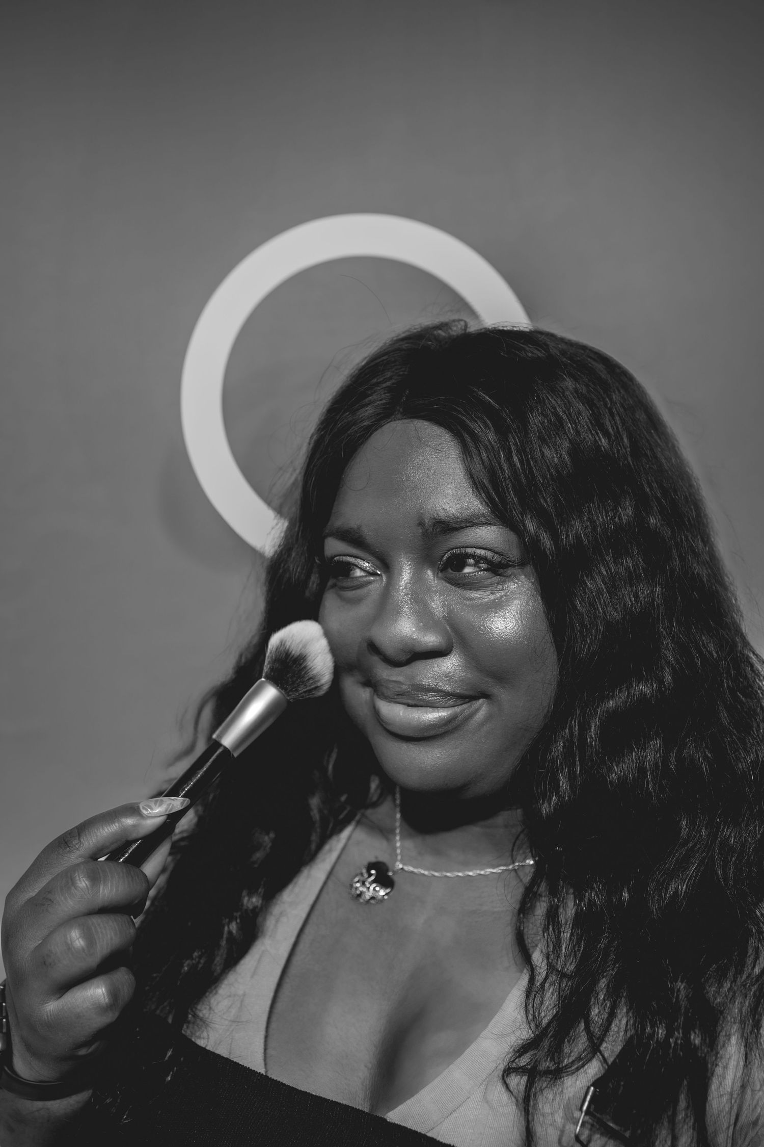 Black and white portrait of a smiling person holding a makeup brush near their face in dramatic lighting.