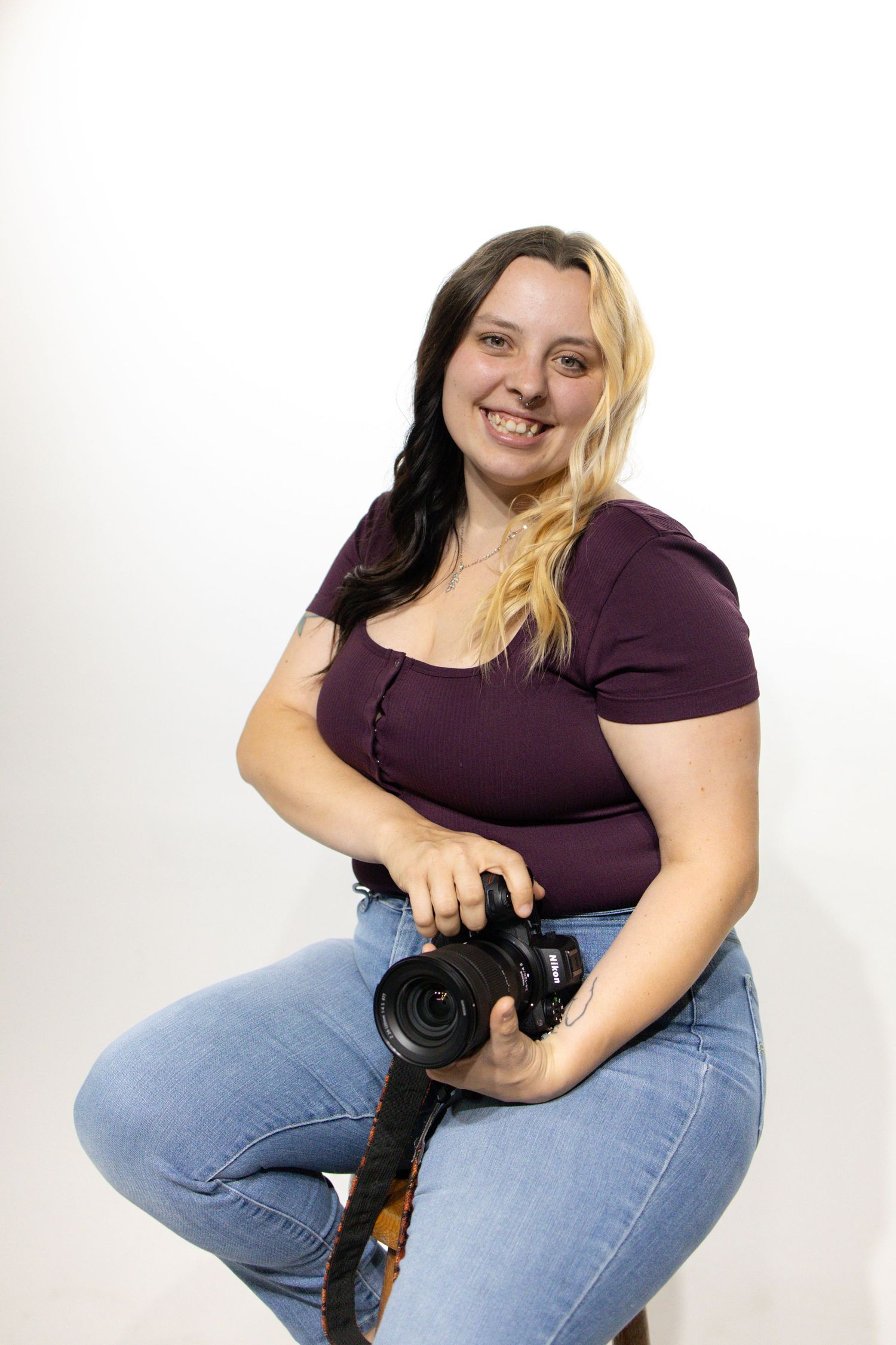 Person in purple shirt and blue jeans holds camera while posing against white background.