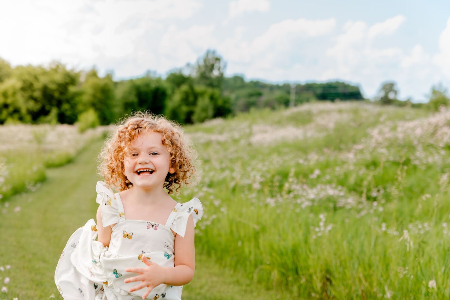 A person in a floral dress stands in a breezy meadow with tall grass and wildflowers on a bright summer day.