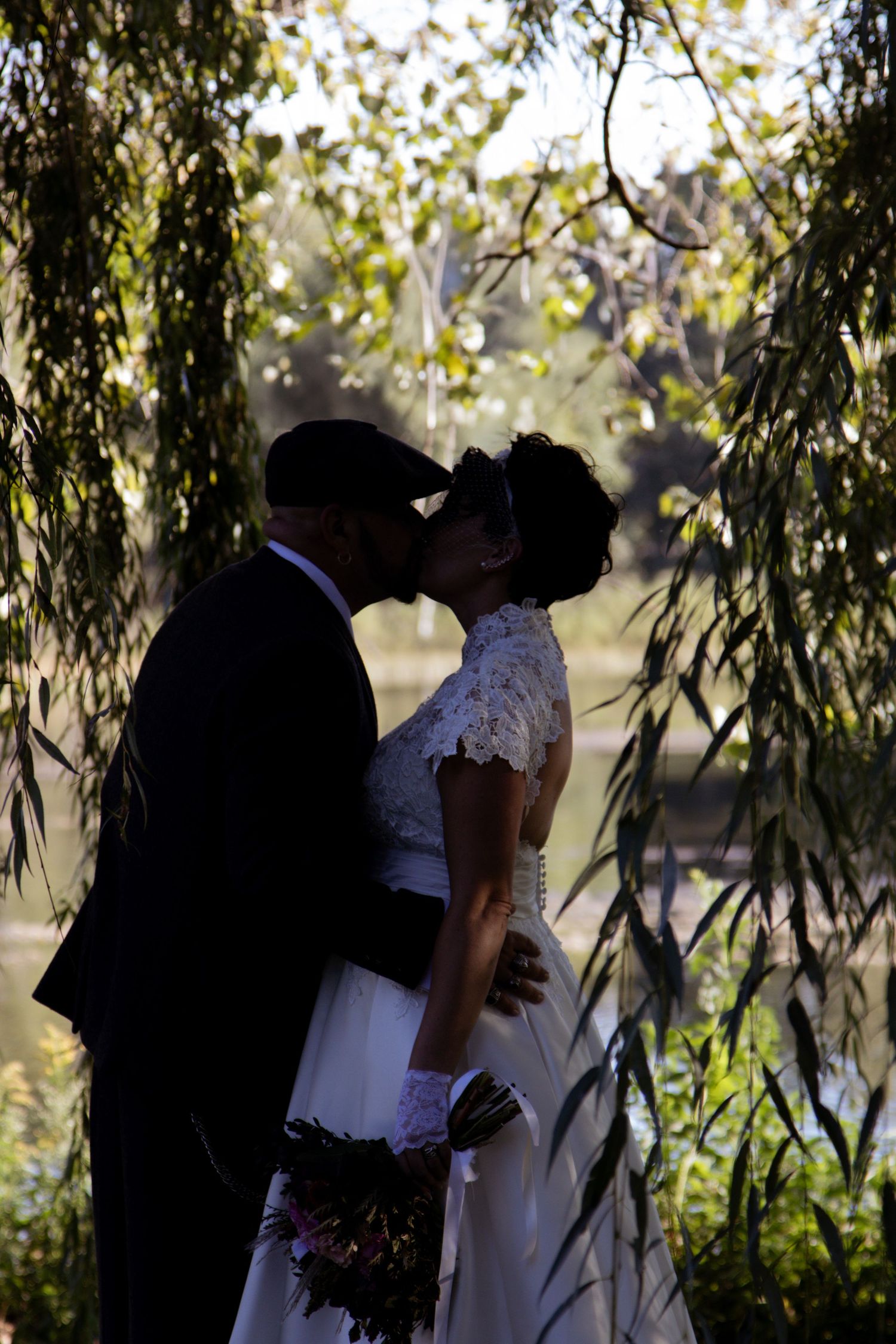 Two figures embrace under willow tree branches in romantic garden setting, silhouetted against natural light.