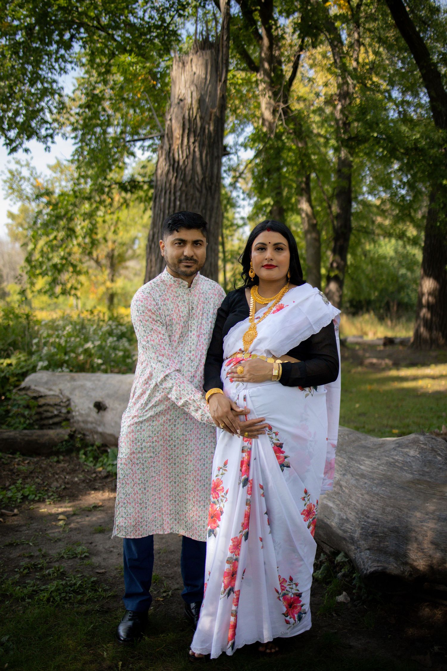 A couple stands near a fallen tree trunk in a sunlit forest setting wearing white and patterned clothing.