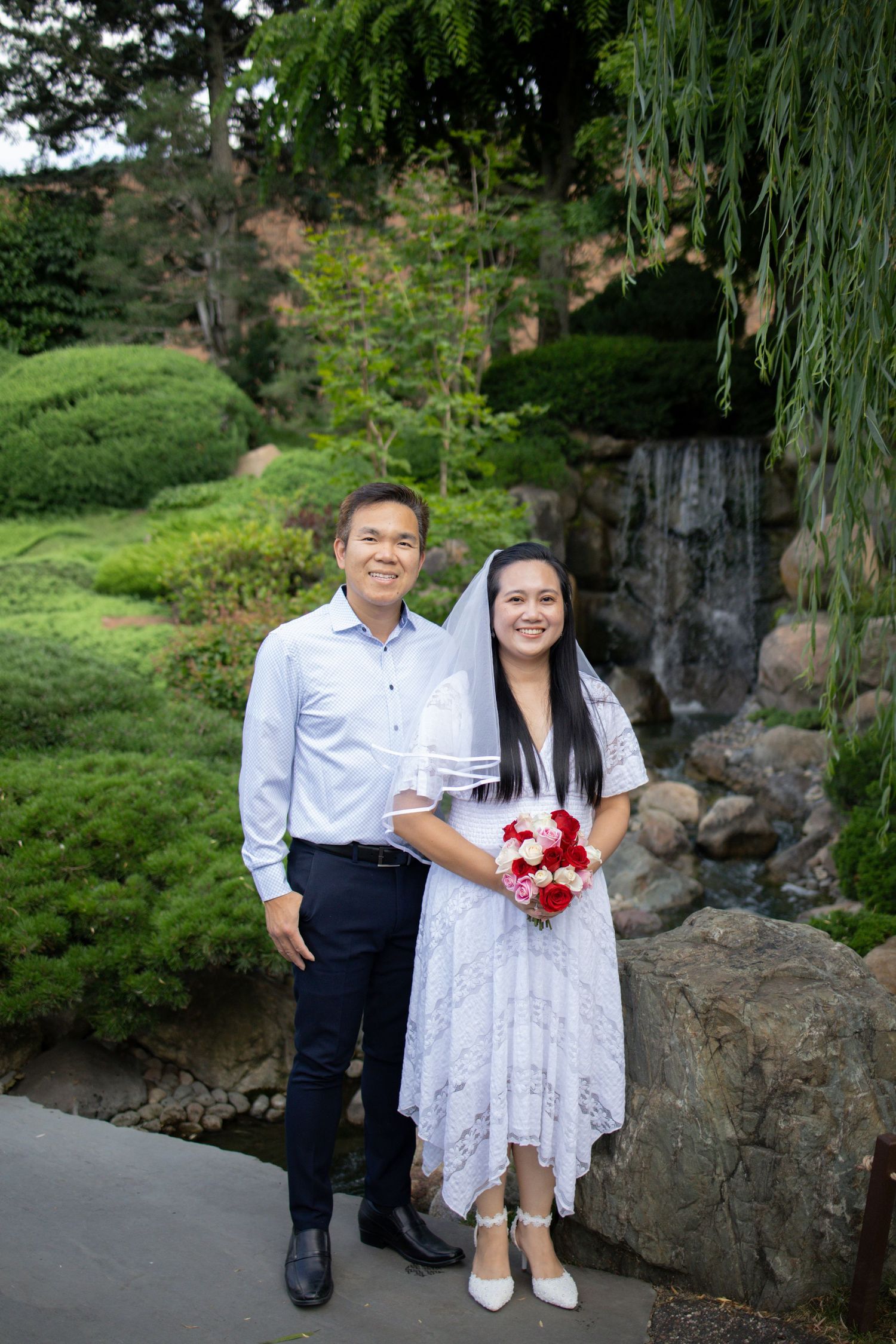 Newlyweds pose together on garden pathway with stone wall and lush greenery in background.