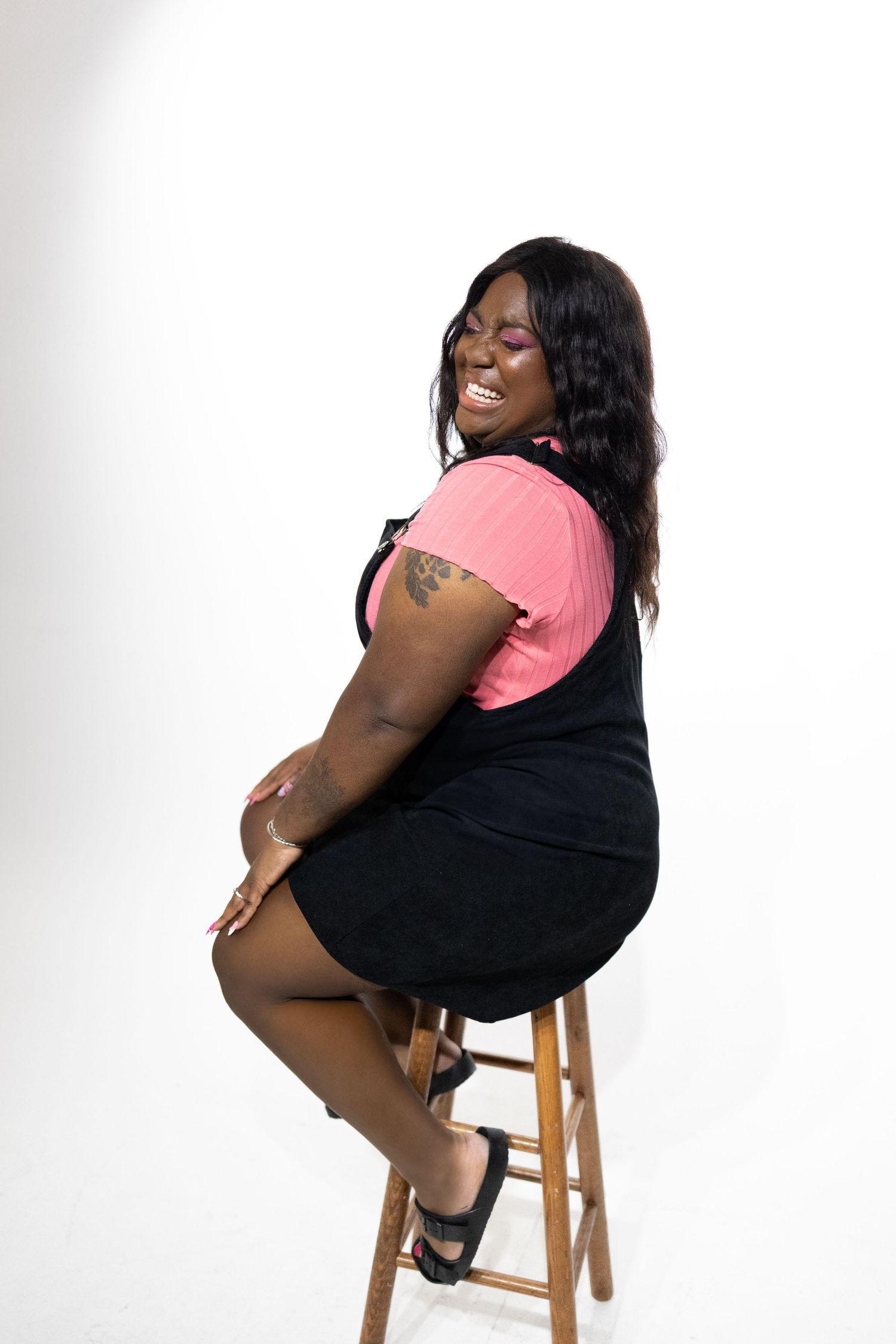 Smiling person wearing pink and black outfit poses on wooden stool against white background.