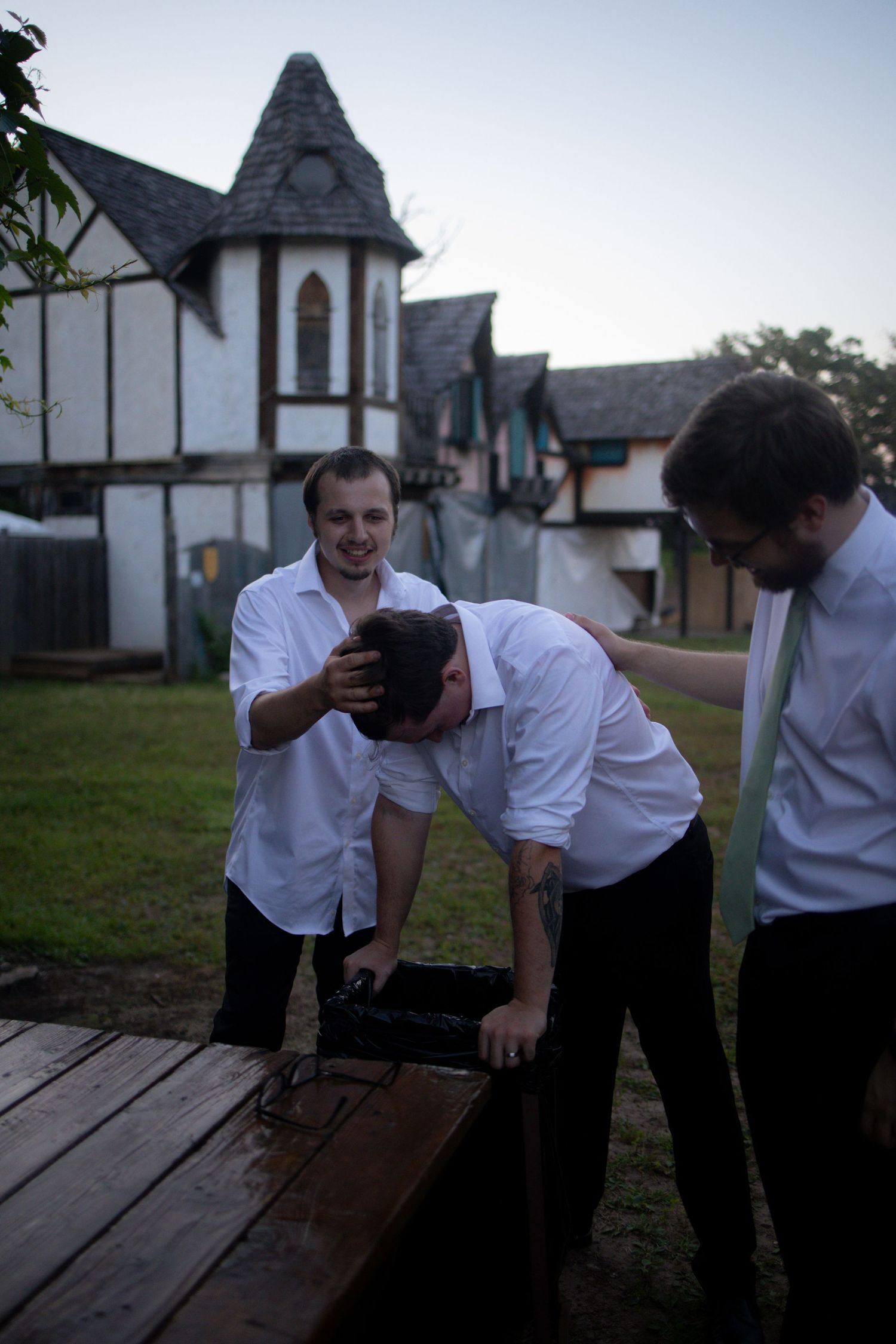 Three people in white shirts gather around a wooden deck with Tudor-style buildings in the background at dusk.