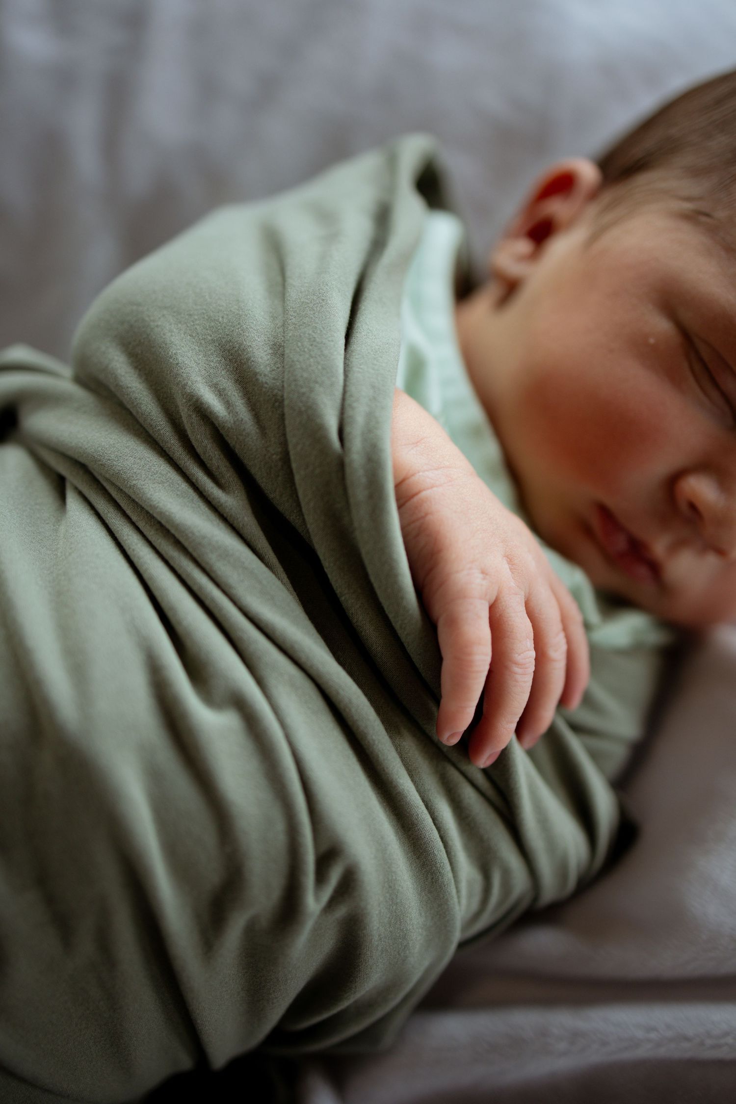 Sleeping baby swaddled in soft mint green blanket rests peacefully on gray bedding.