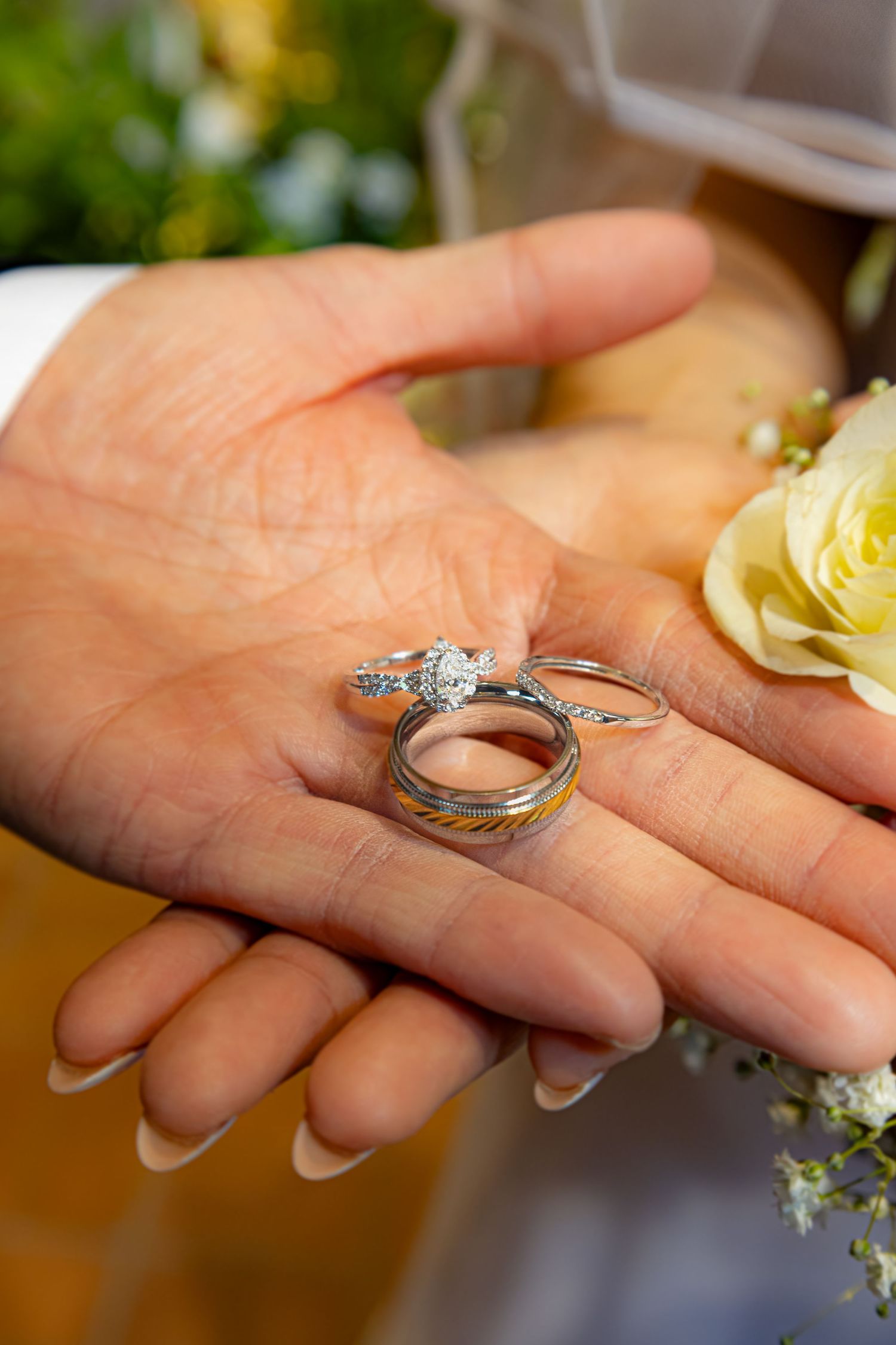 A hand holds two wedding rings against a soft backdrop of yellow roses and white flowers.