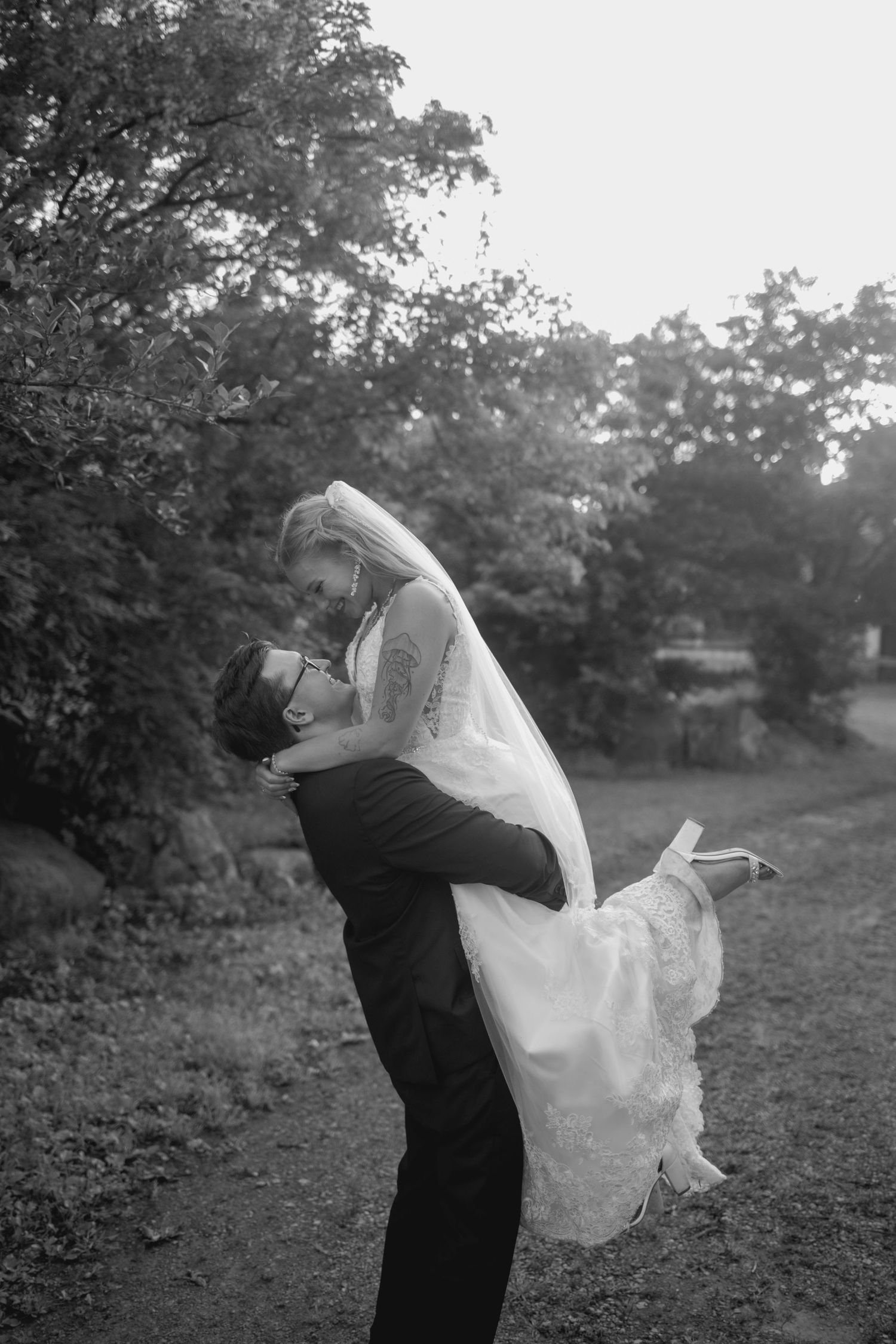 A romantic black and white wedding photograph of a couple embracing outdoors, with the bride being lifted in her flowing dress.