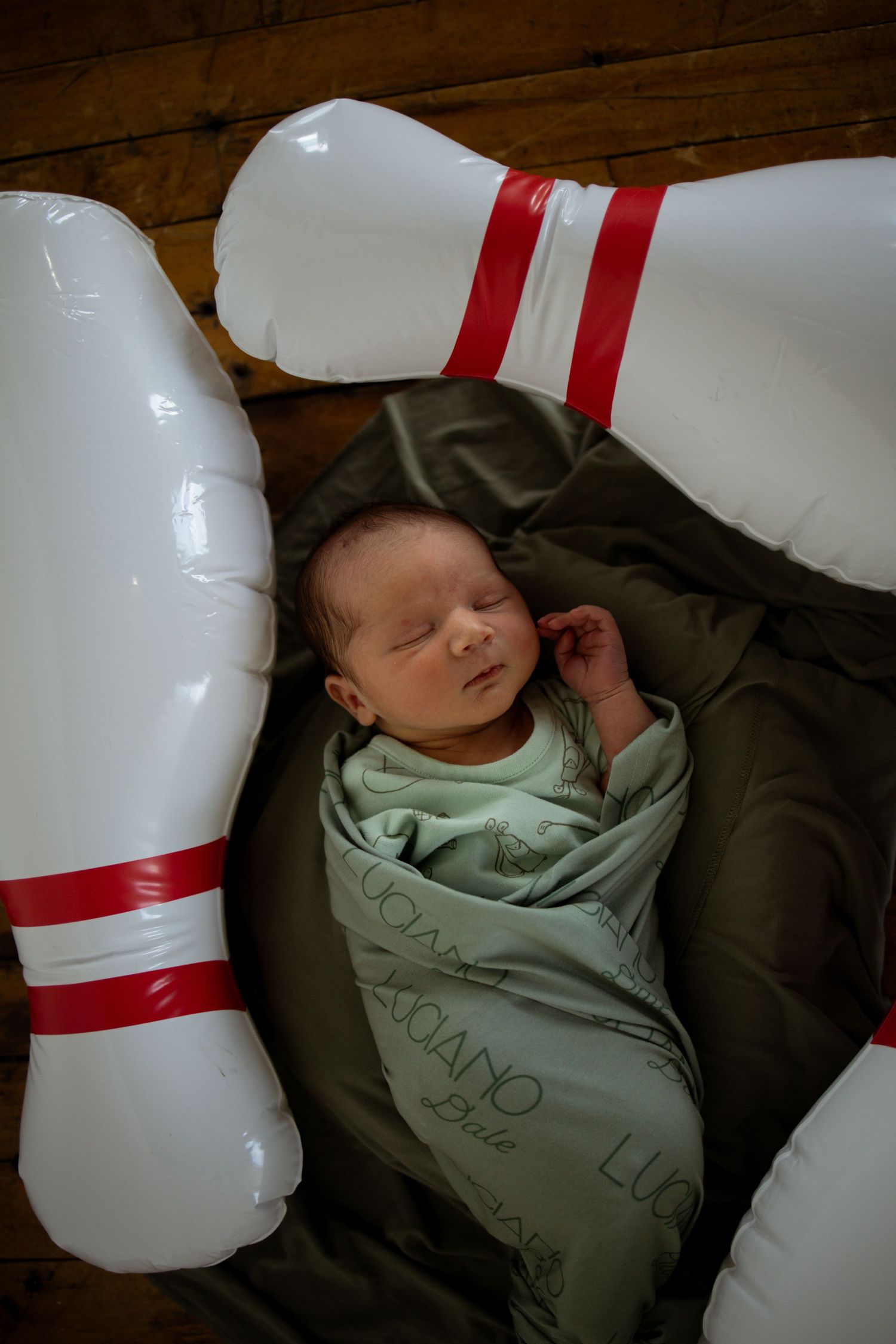 Sleeping baby wrapped in mint green blanket lying between large white bowling pins with red stripes.